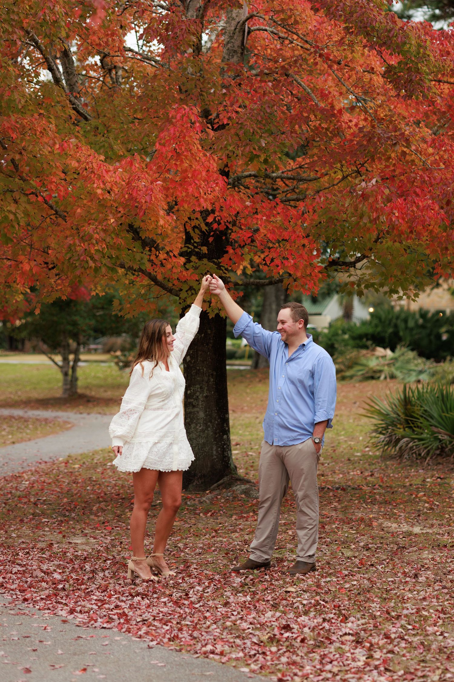 A couple shares playful moments under vibrant red autumn trees with fallen leaves scattered on the ground.