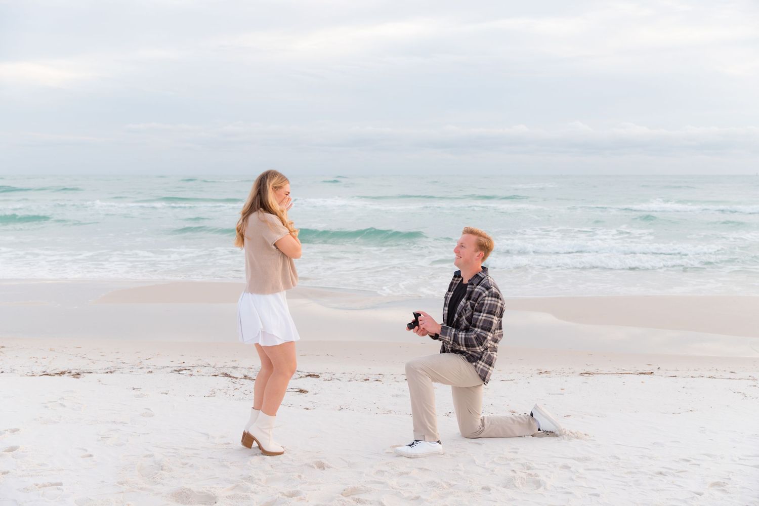 A surprise beach proposal unfolds as someone kneels on white sand with ocean waves behind them.