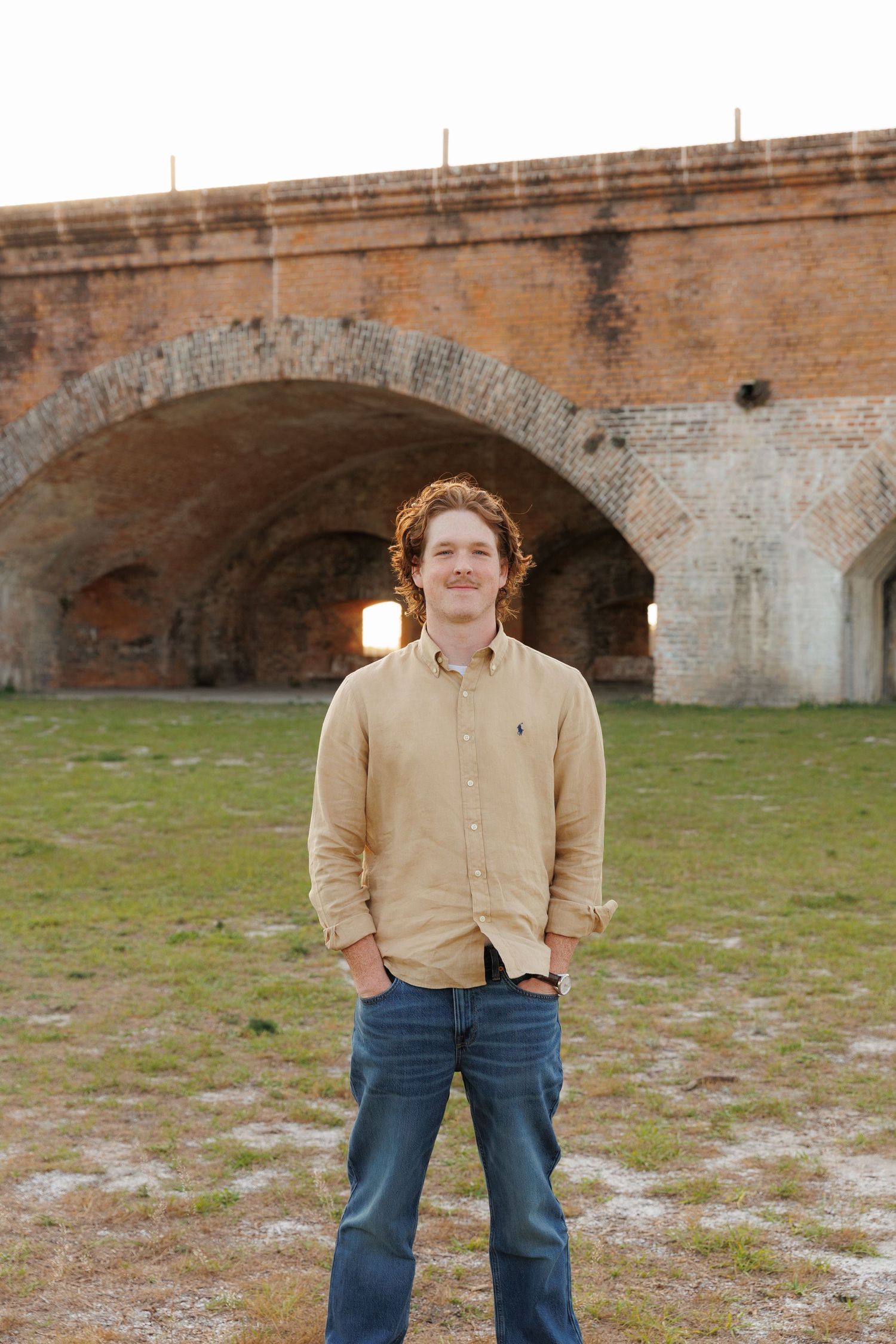 Young man with curly red hair in beige Polo shirt and jeans smiling outdoors near historic brick arched structure.
