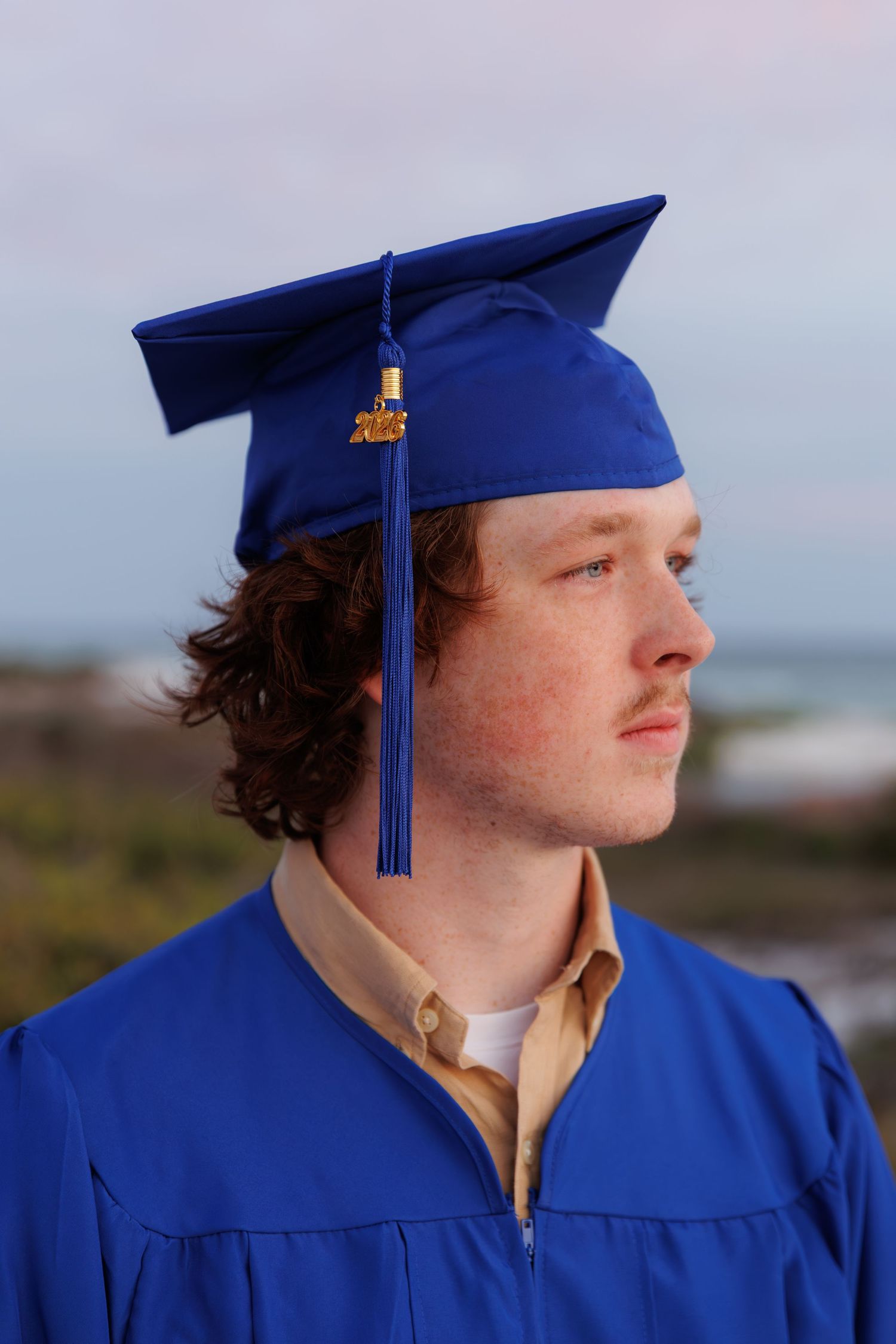 Young man in blue graduation cap and gown poses outdoors near ocean, looking thoughtfully into the distance.