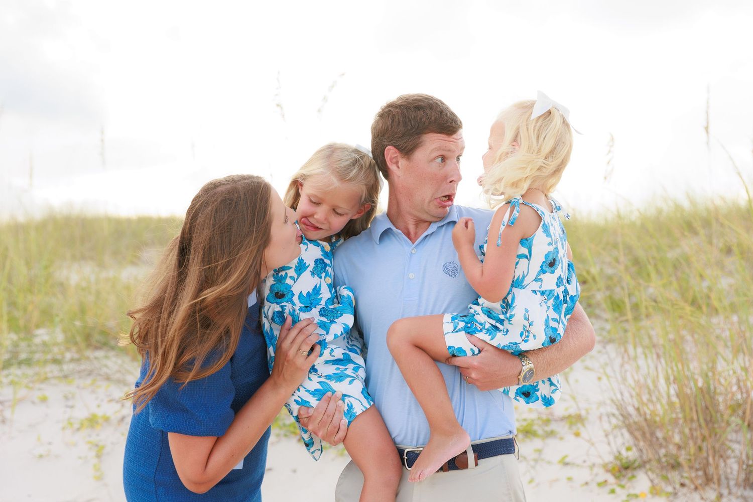 Family of four enjoying a beach photoshoot, parents holding two young girls in matching floral outfits on white sand.