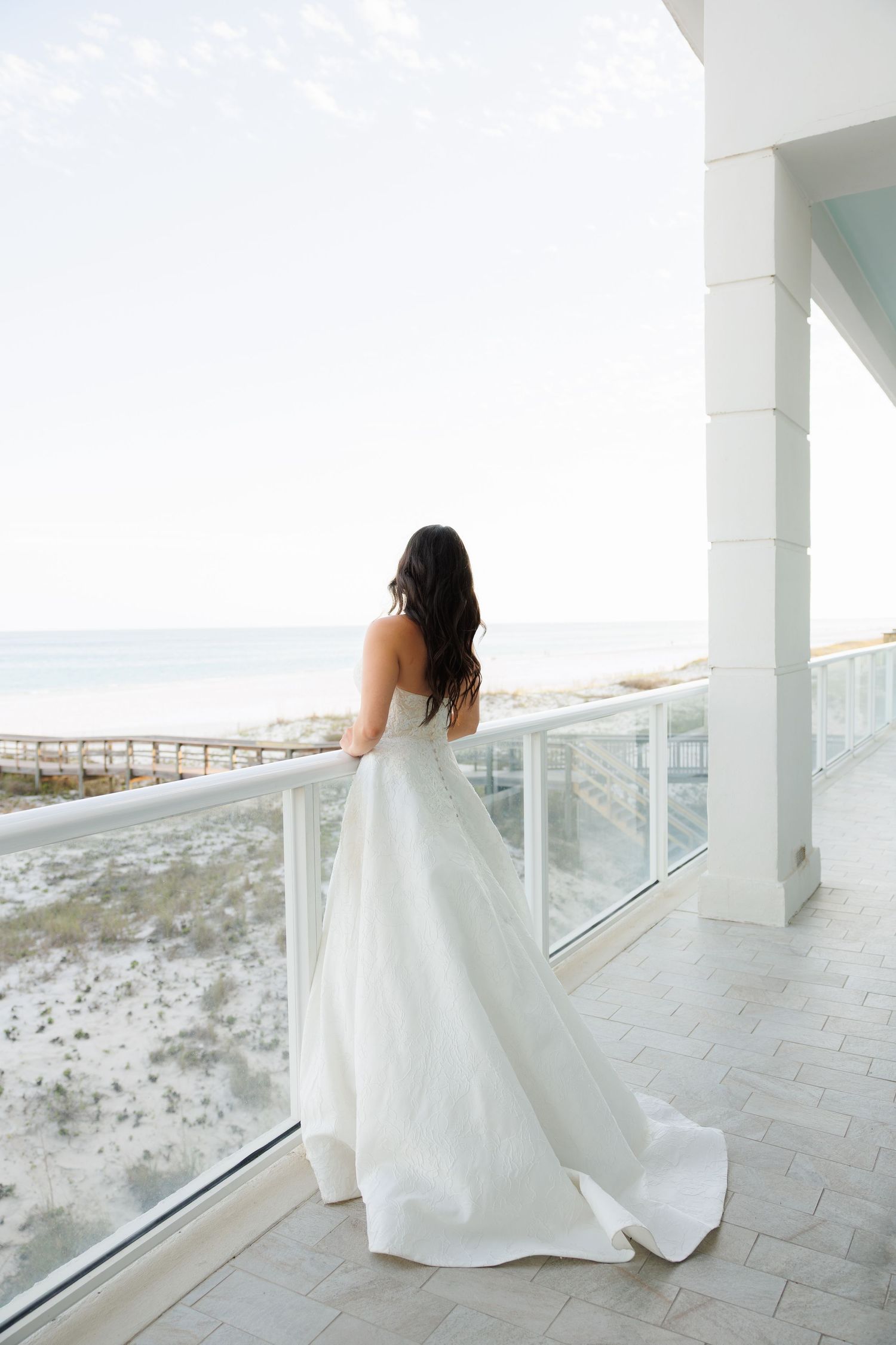 Bride in strapless white ball gown stands on beachfront balcony overlooking sandy shore and ocean.