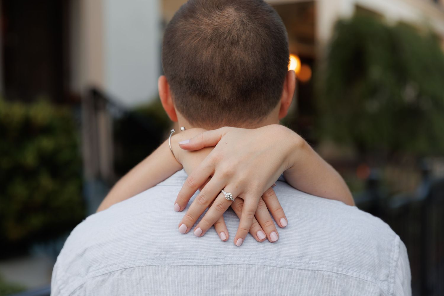Couple embracing from behind, woman's hand with engagement ring resting on man's shoulder outdoors.