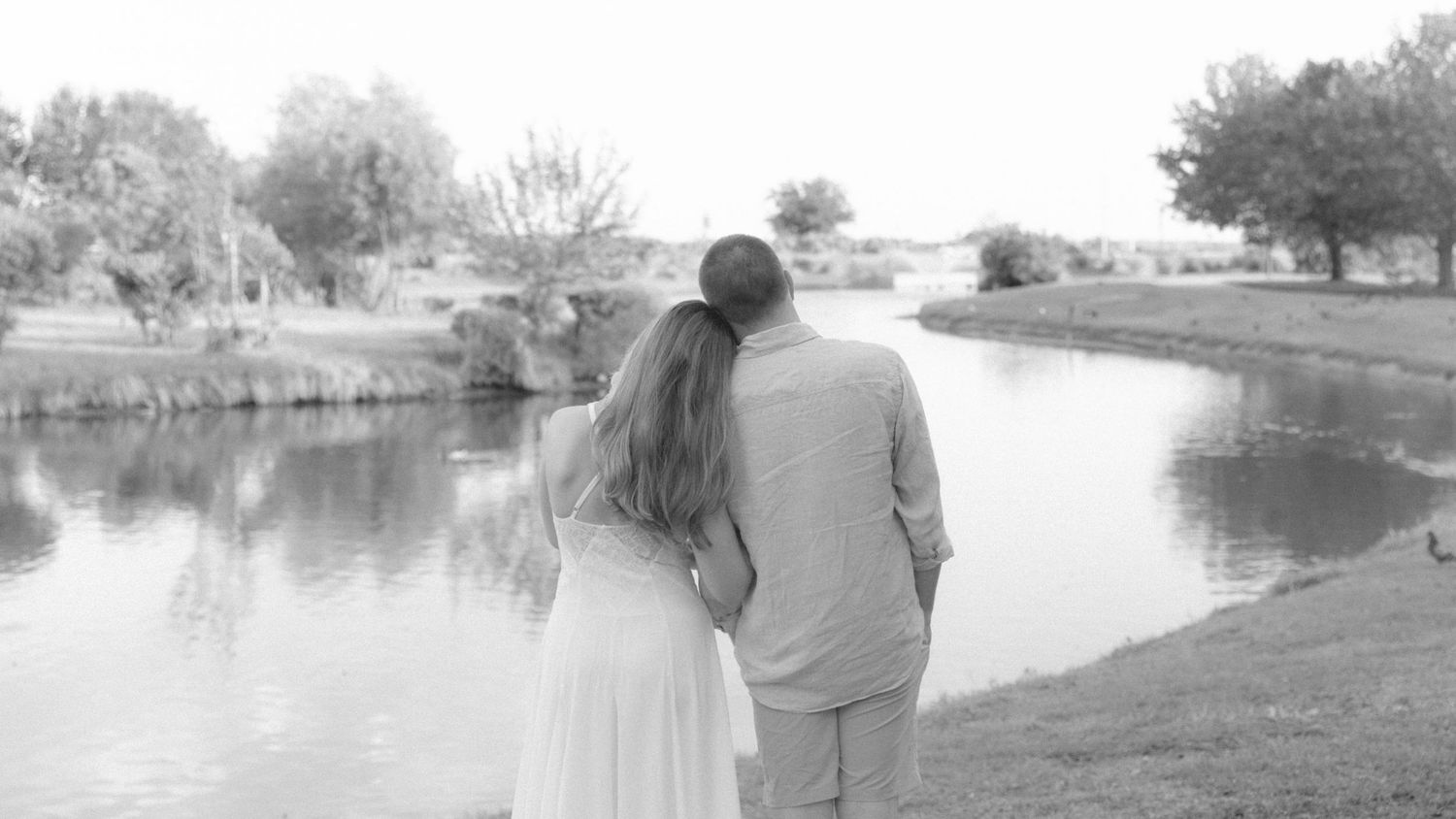 Black and white photo of a couple standing by a serene pond, viewed from behind on their wedding day.