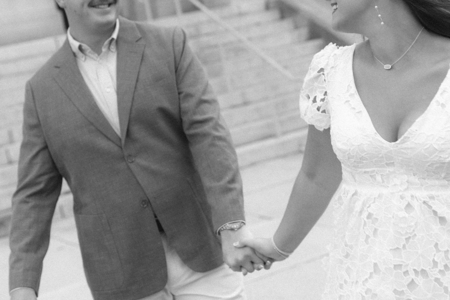 Black and white photo of a couple holding hands, man in gray suit and woman in white lace wedding dress.