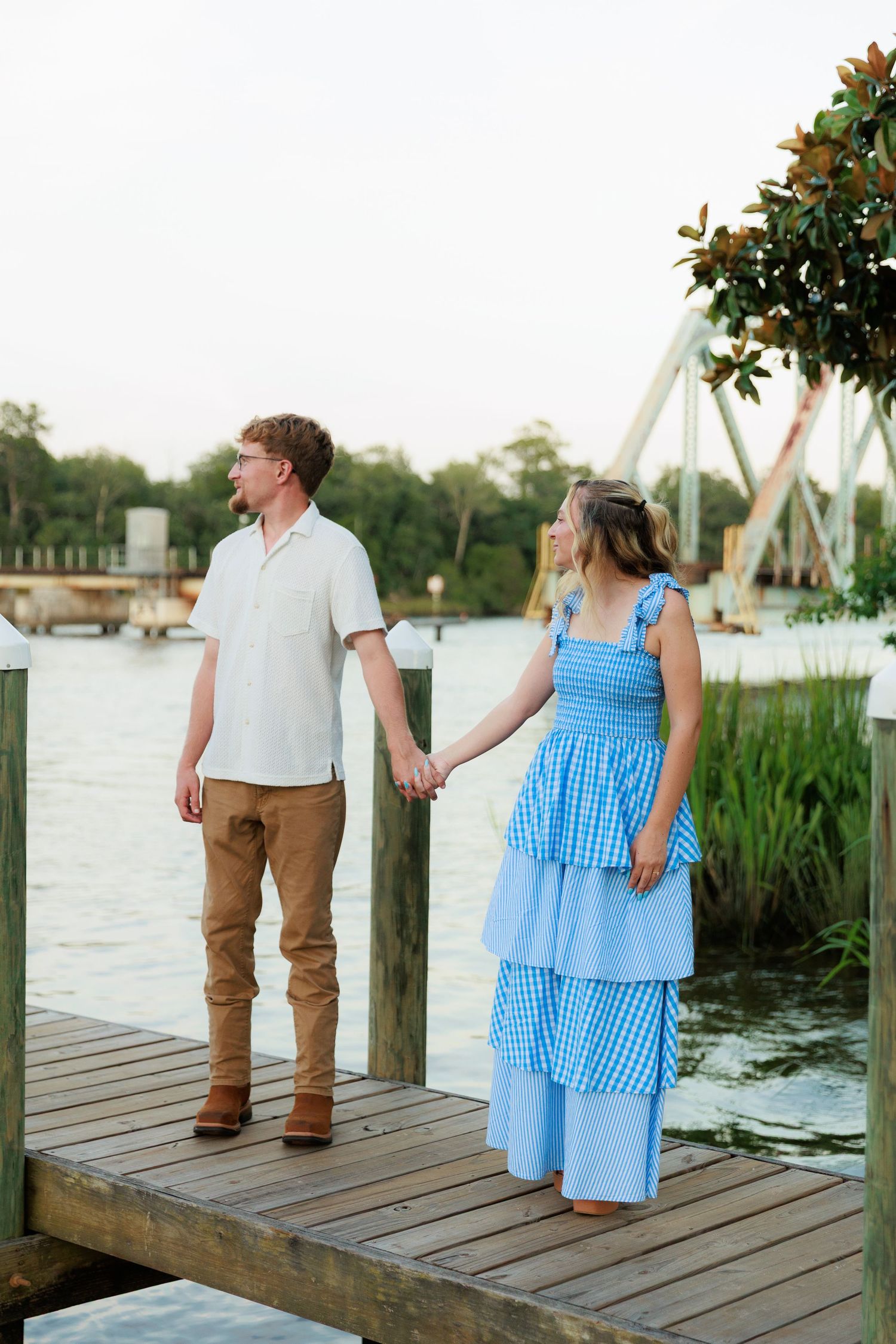 Couple holding hands on a wooden dock by the water, woman in blue gingham tiered dress, man in white linen shirt.