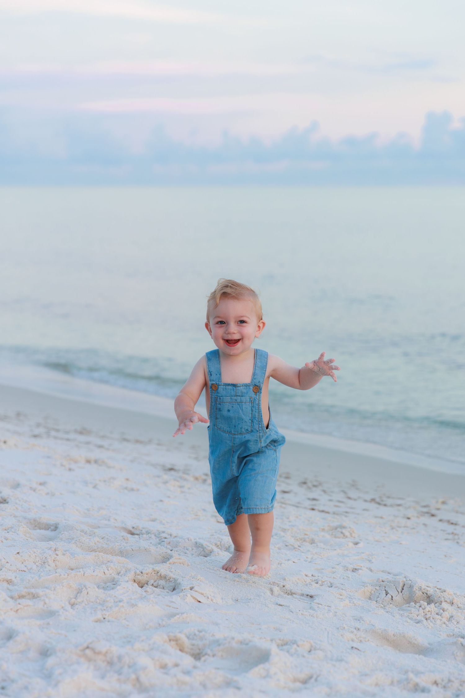 Happy toddler in denim overalls laughing and running barefoot on a white sandy beach at sunset.