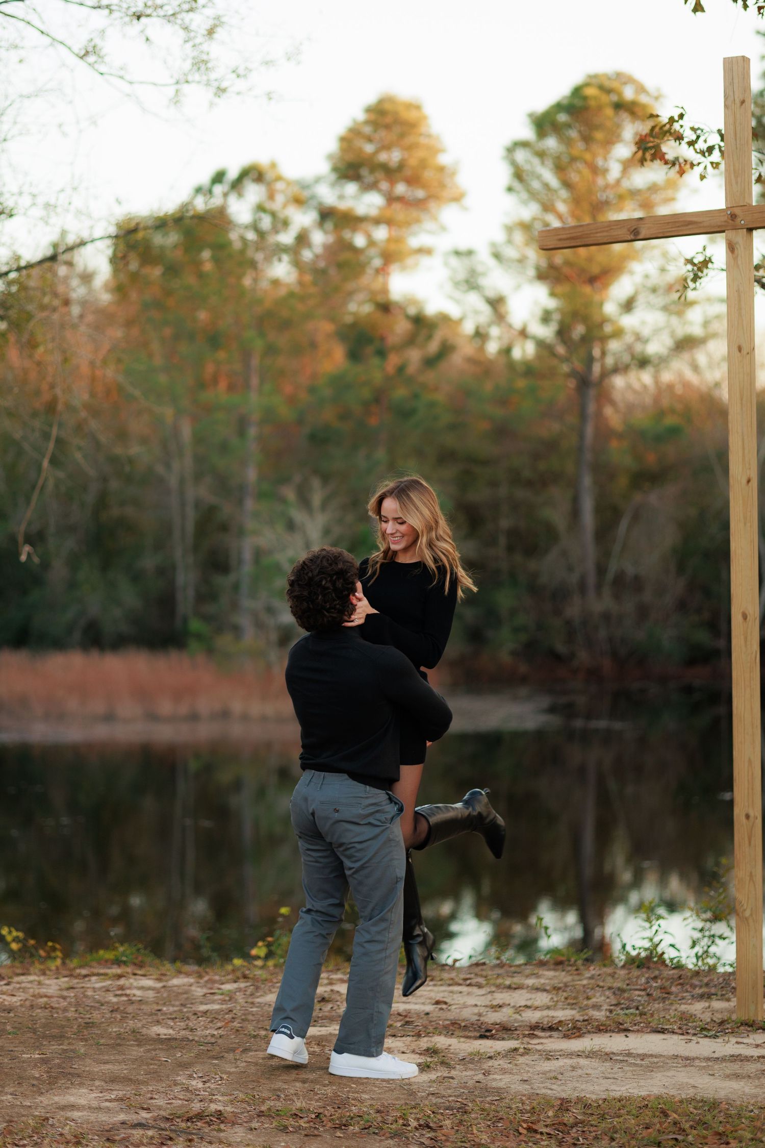 A couple shares a playful moment by a serene lake during autumn with trees in the background.