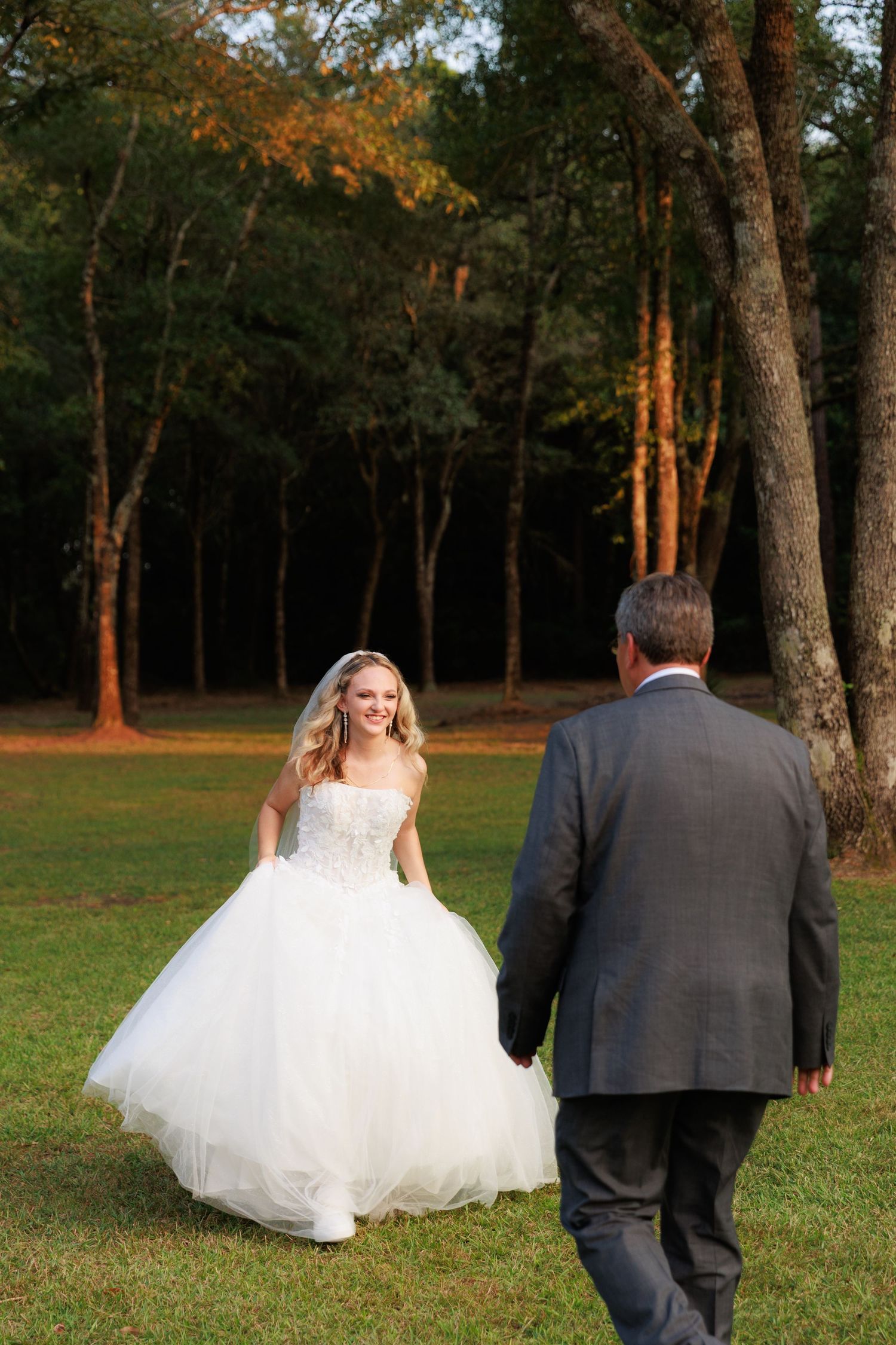 A bride in a flowing white dress walks with a man in a dark suit through a sunlit wooded area.