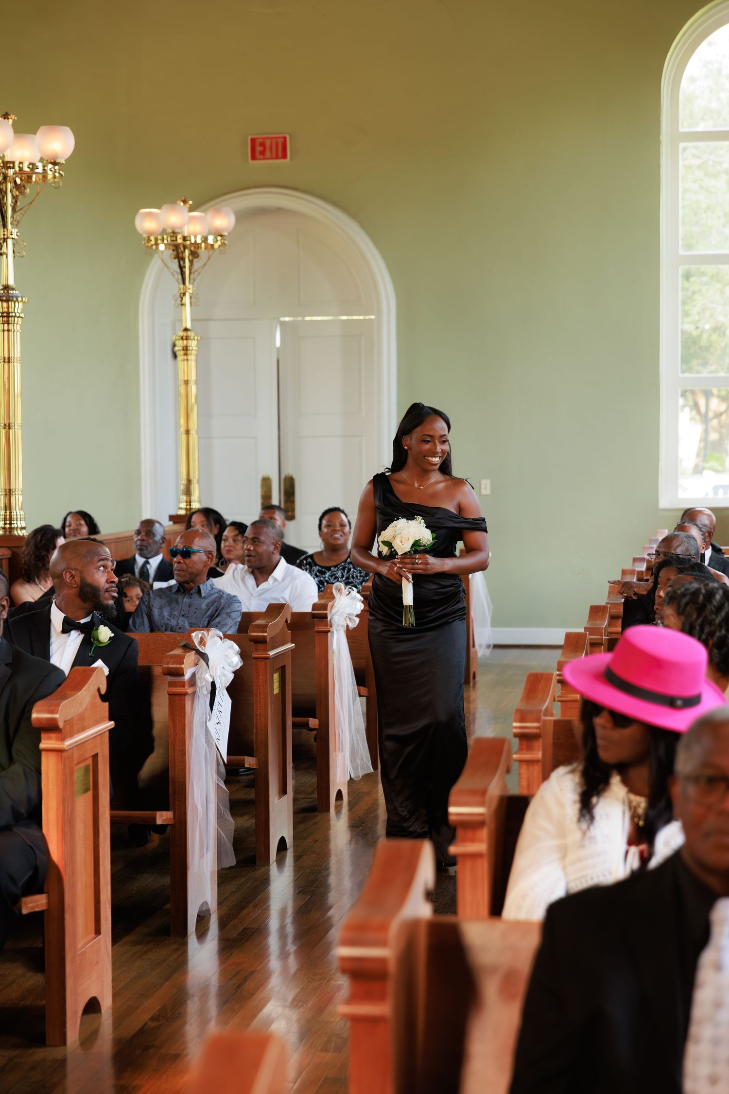 Wedding ceremony guests stand in church pews decorated with white bows and elegant chandeliers overhead.