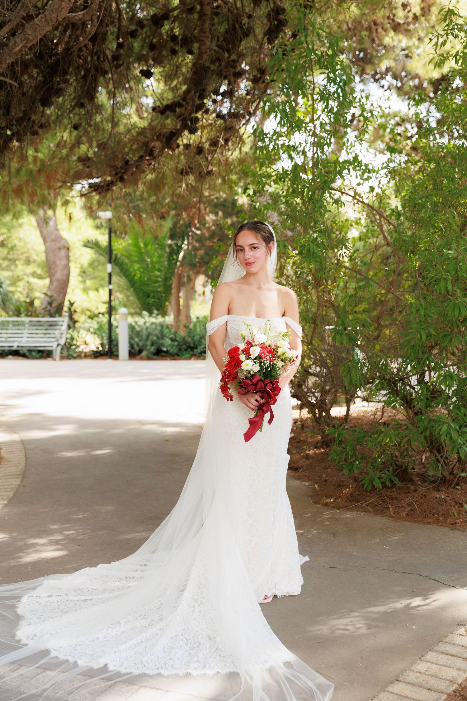 A bride in a flowing white lace gown poses gracefully in a garden setting with sunlight filtering through trees.