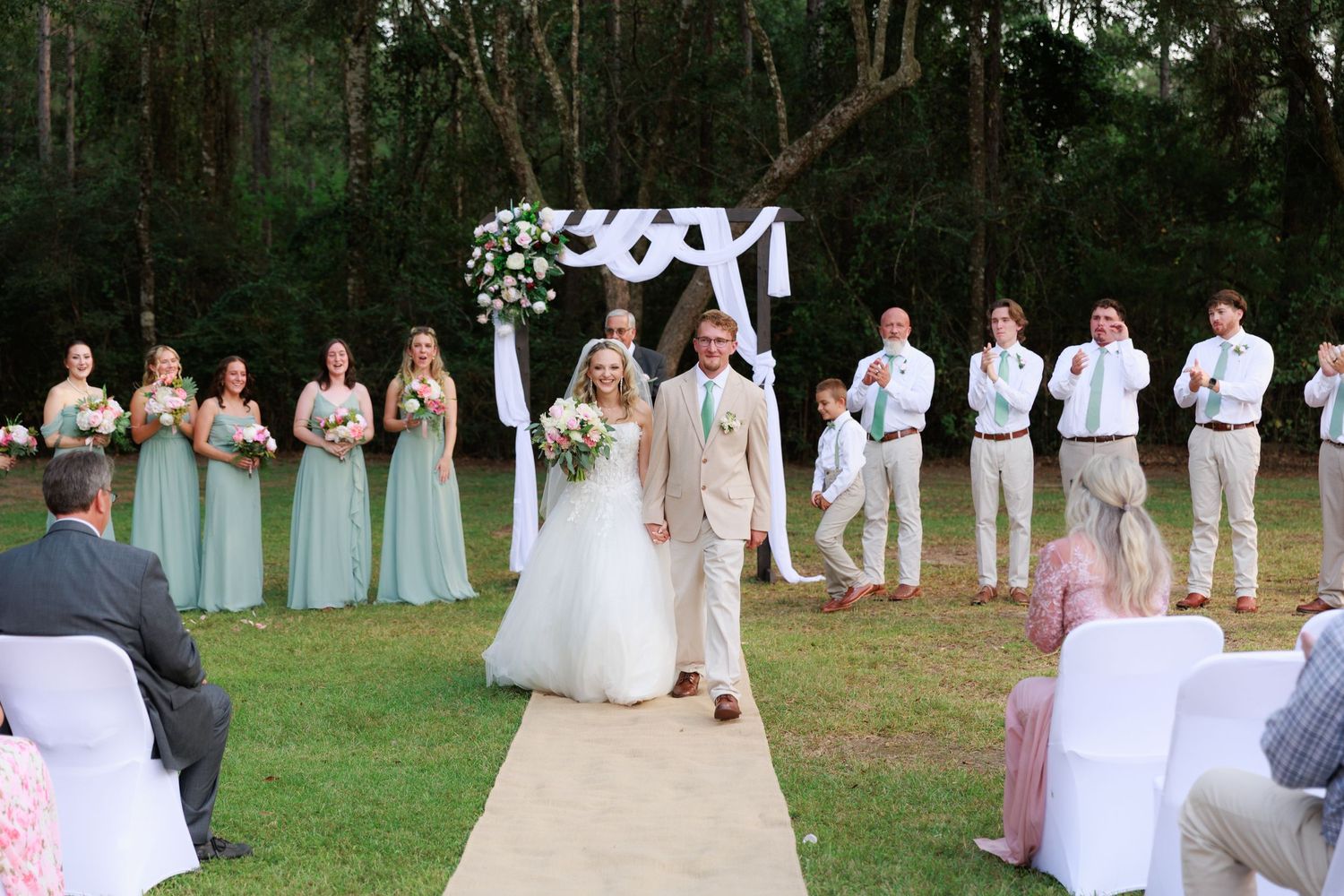 A wedding party in light blue and white attire poses together on a grassy lawn for formal photographs.