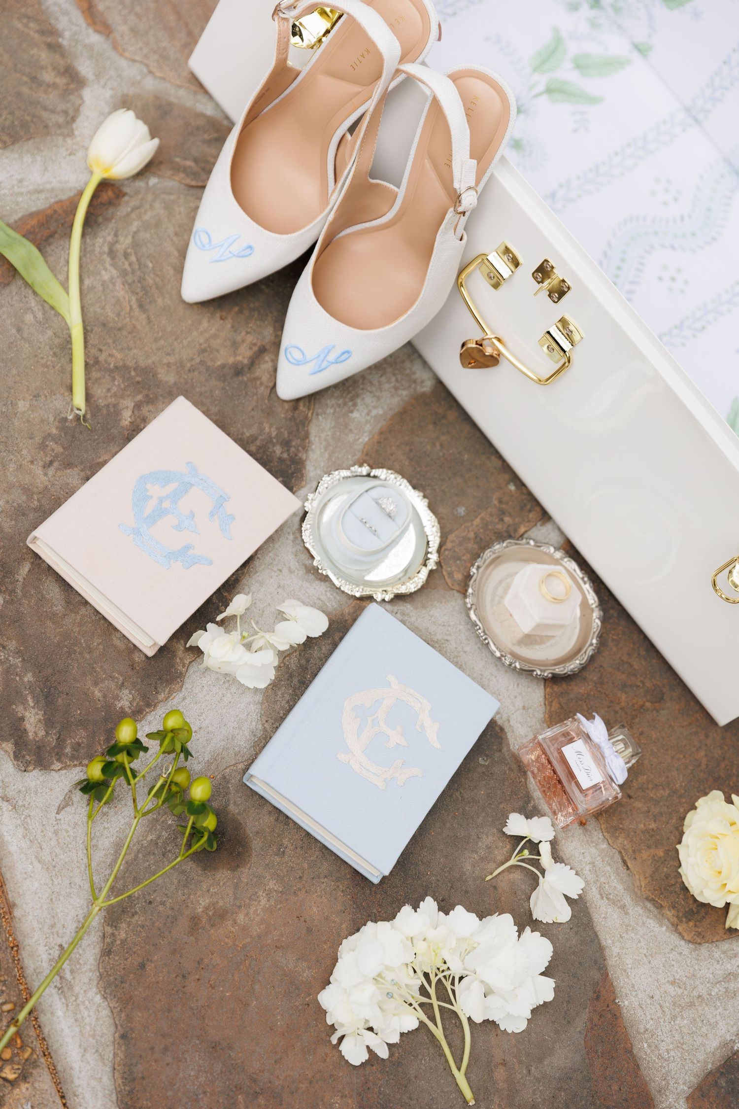 Elegant bridal flat lay with monogrammed white heels, perfume, compact mirror, and white flowers on stone surface.