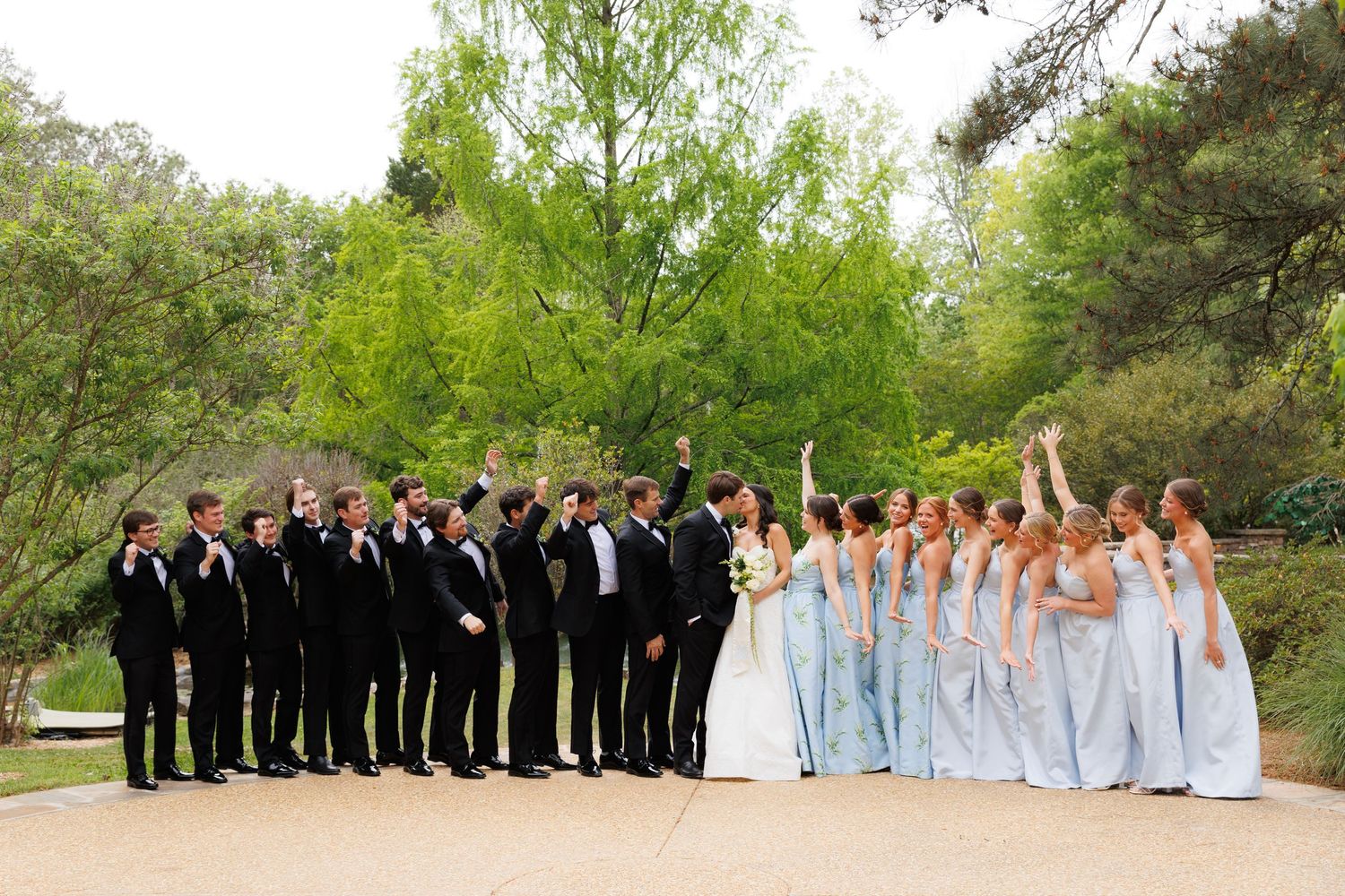 Wedding party celebrates outdoors, bridesmaids in light blue gowns and groomsmen in black tuxedos raising arms joyfully.