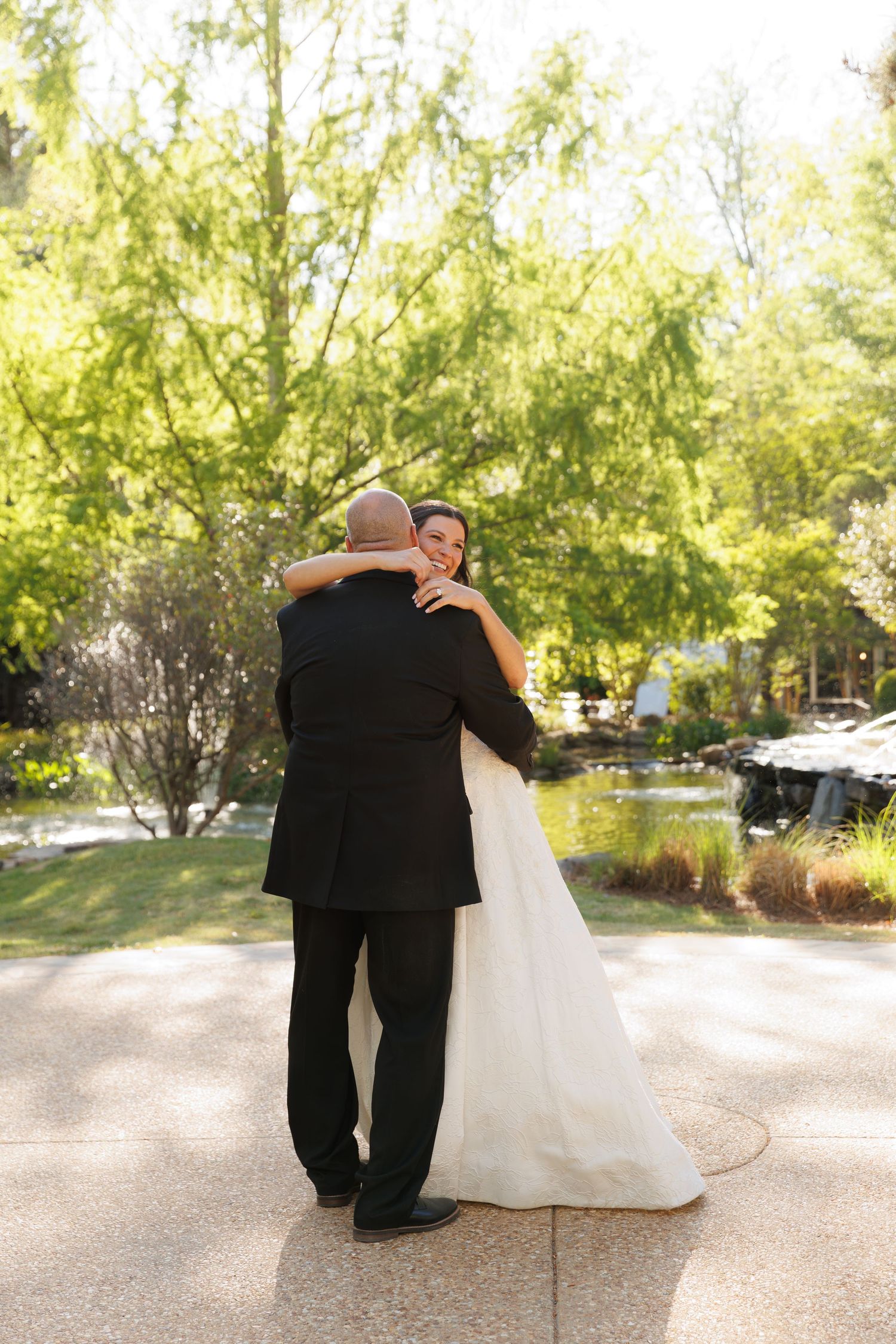 Smiling bride embraces groom in a warm hug outdoors surrounded by lush green trees on their wedding day.