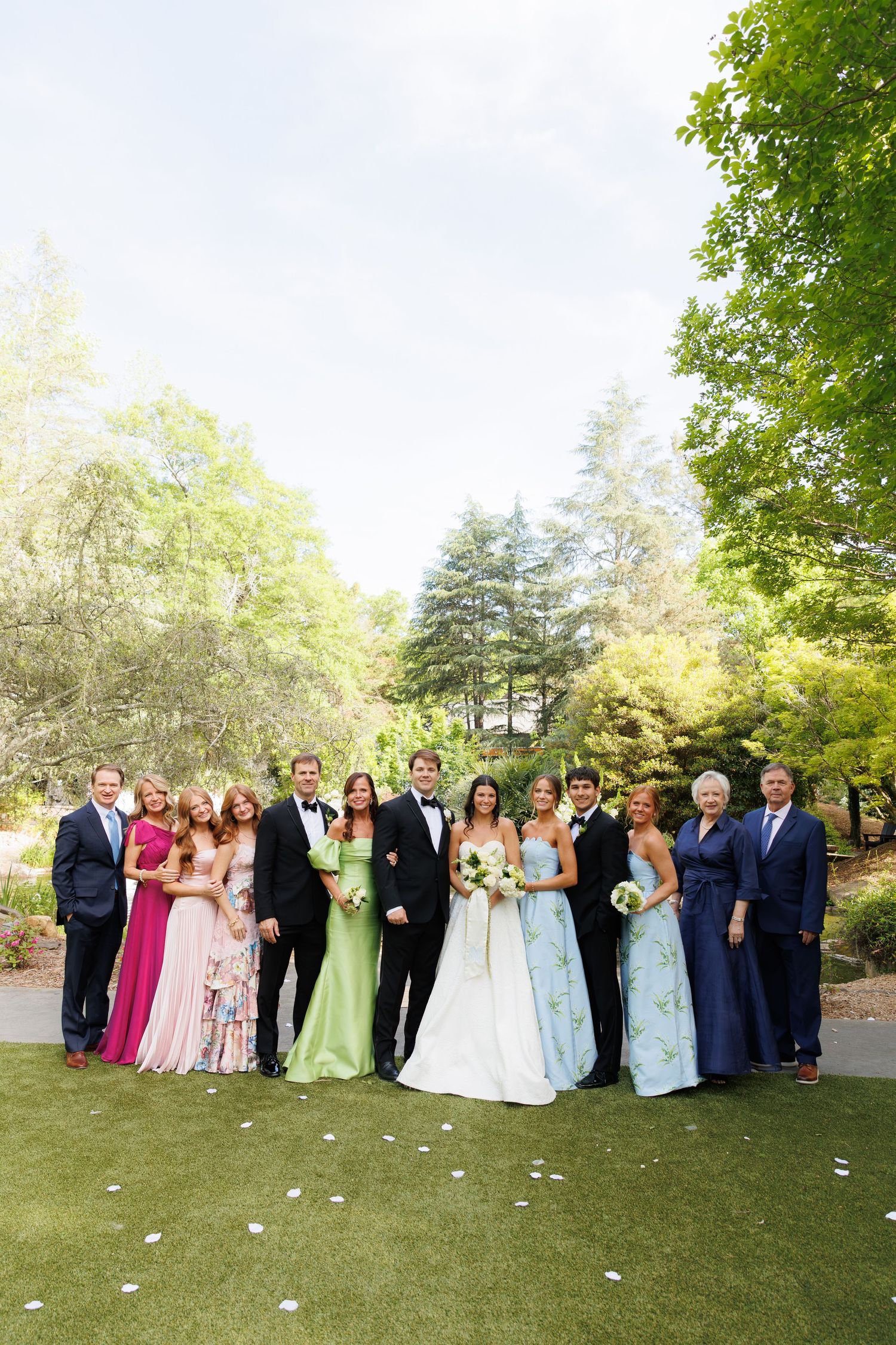Wedding party poses outdoors on green lawn surrounded by lush trees, bride in white gown with floral wedding party.