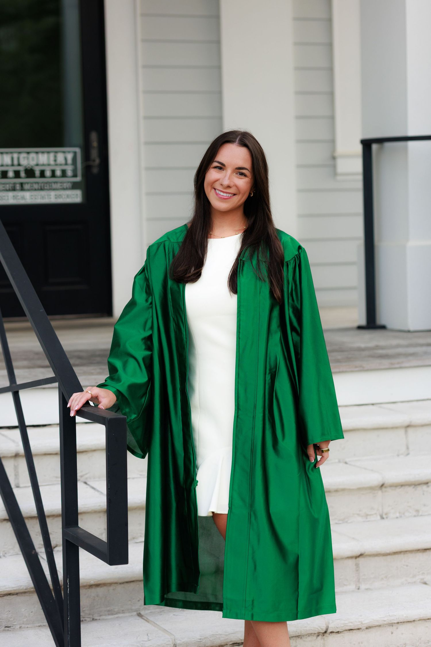Smiling young woman in green graduation gown over white dress, standing on white steps outside a building.