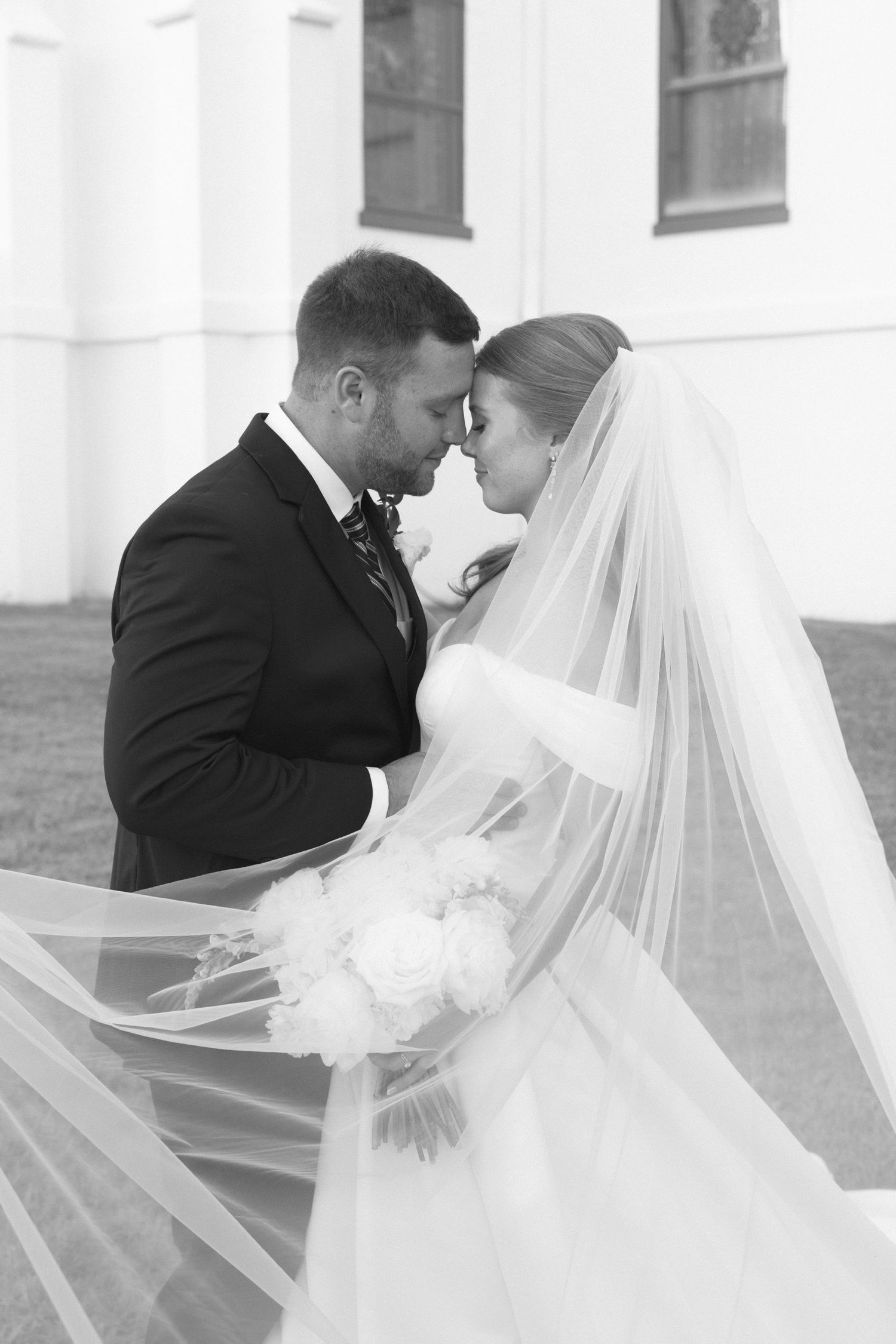 Black and white portrait of bride and groom touching foreheads tenderly as veil flows around them.