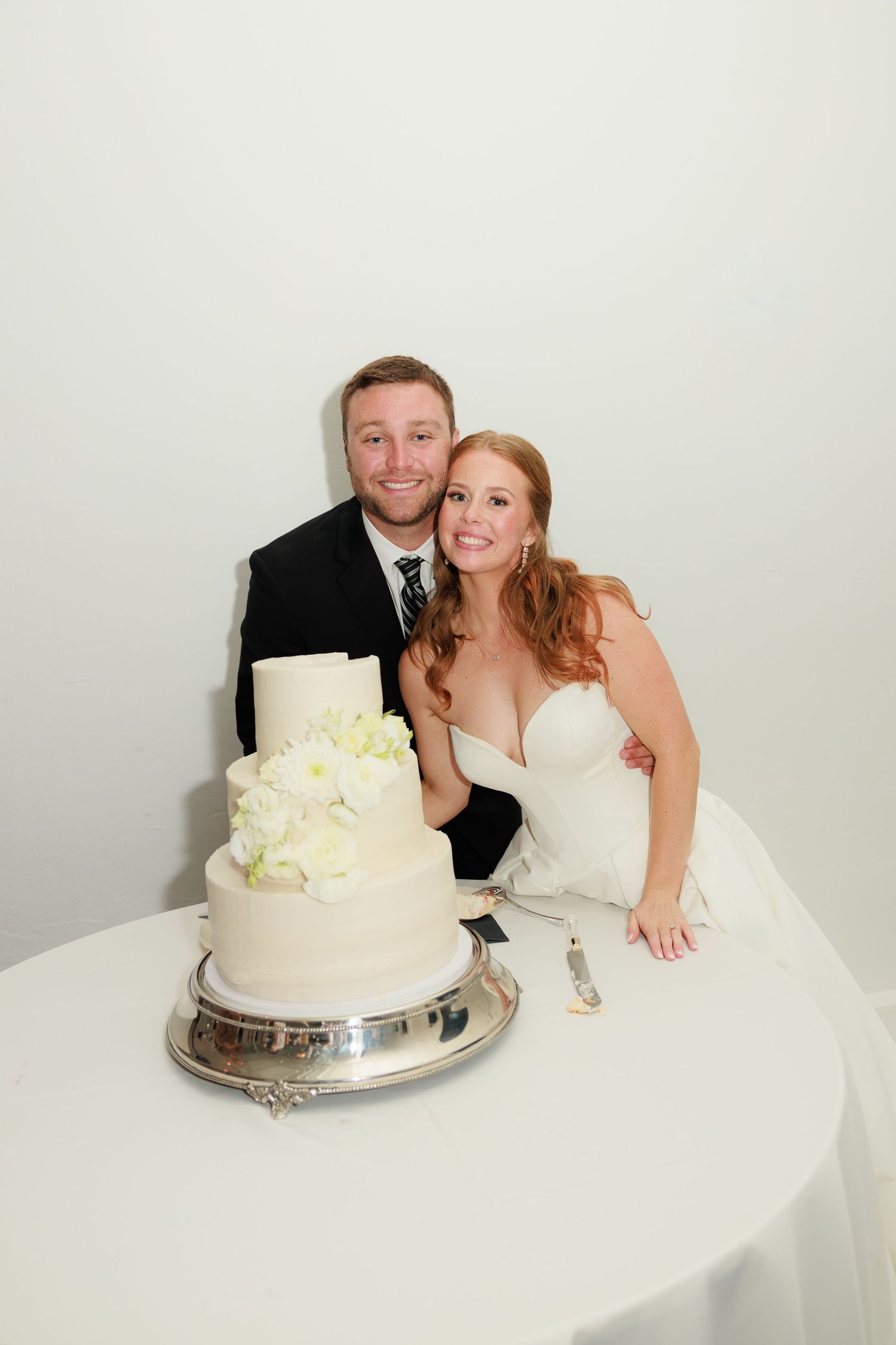 Bride and groom smiling together behind white tiered wedding cake with floral decor.