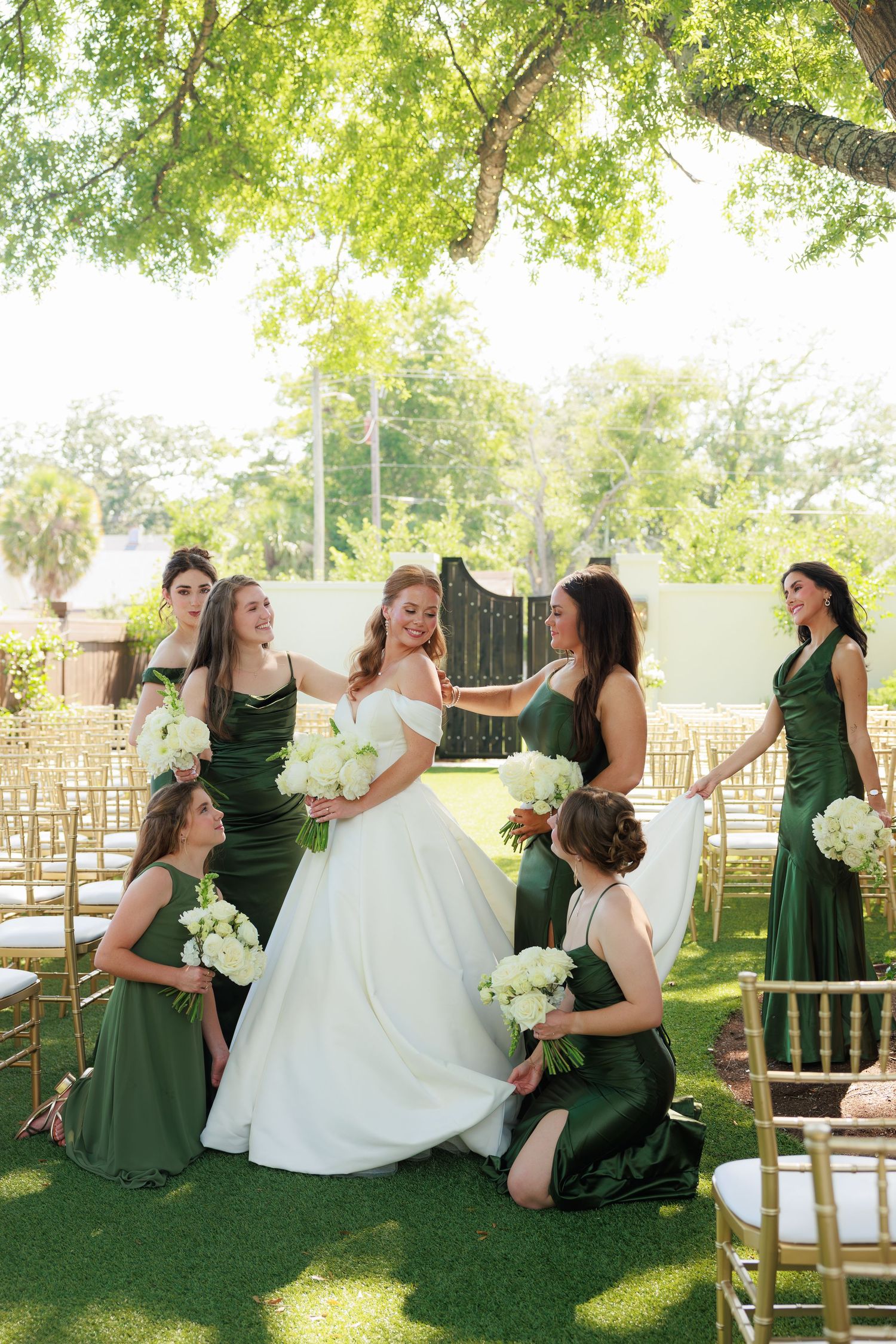 Bride in white off-shoulder gown surrounded by bridesmaids in green satin dresses holding white bouquets at outdoor wedding.