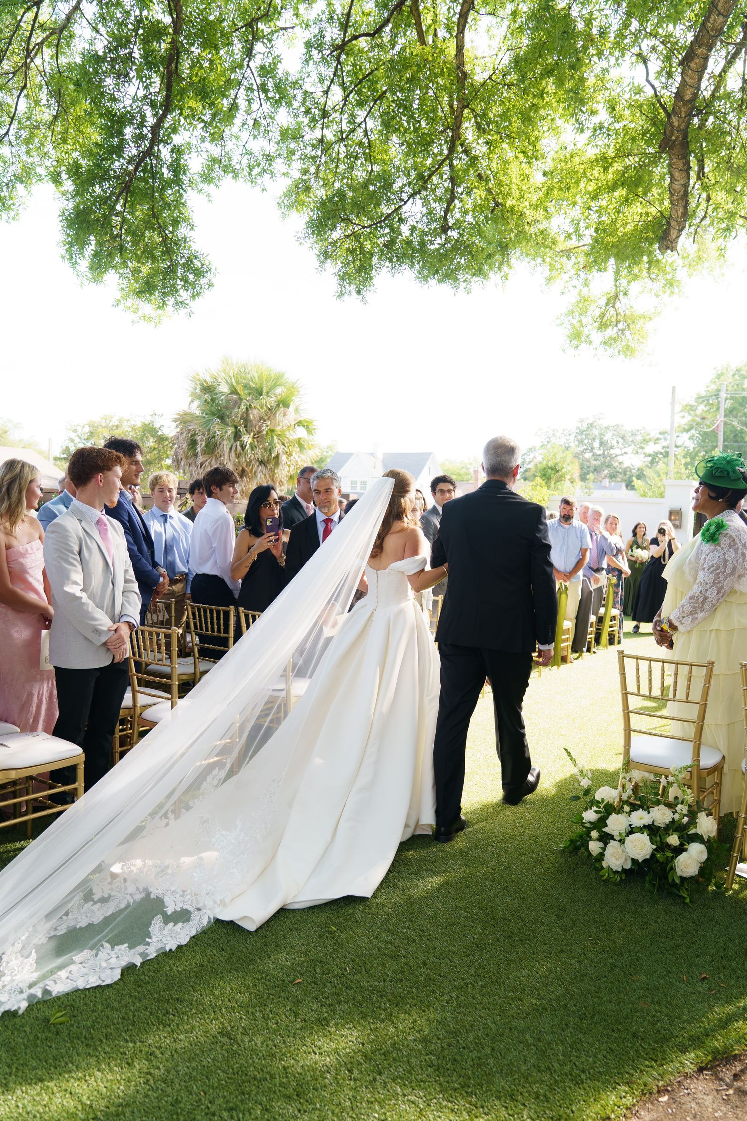 Bride in elegant white gown with long lace veil walks down outdoor aisle with guests seated on gold chairs.