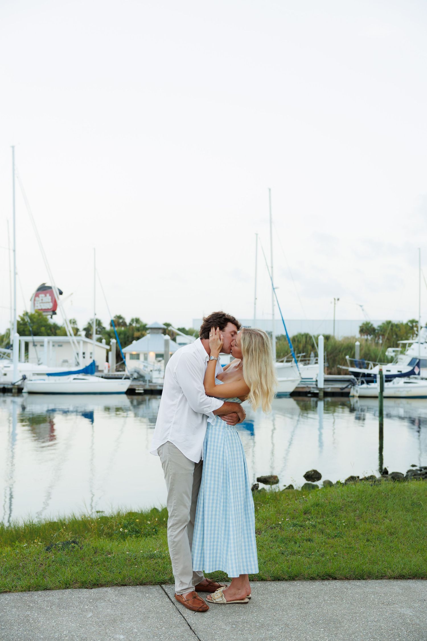 Romantic couple shares a kiss by a peaceful marina with sailboats in the background.
