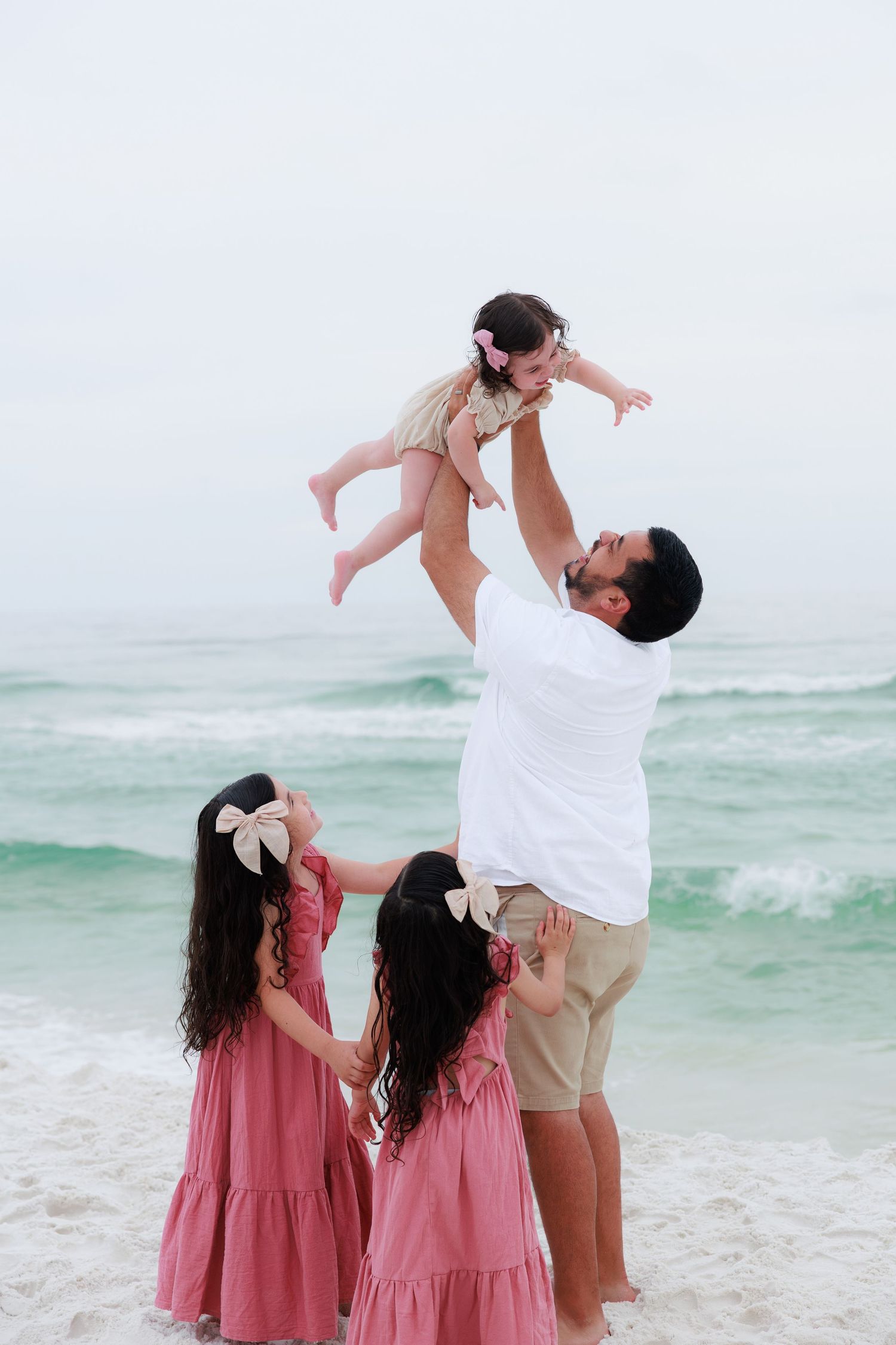 Father lifting baby girl in air at beach while two daughters in pink dresses watch the ocean waves.