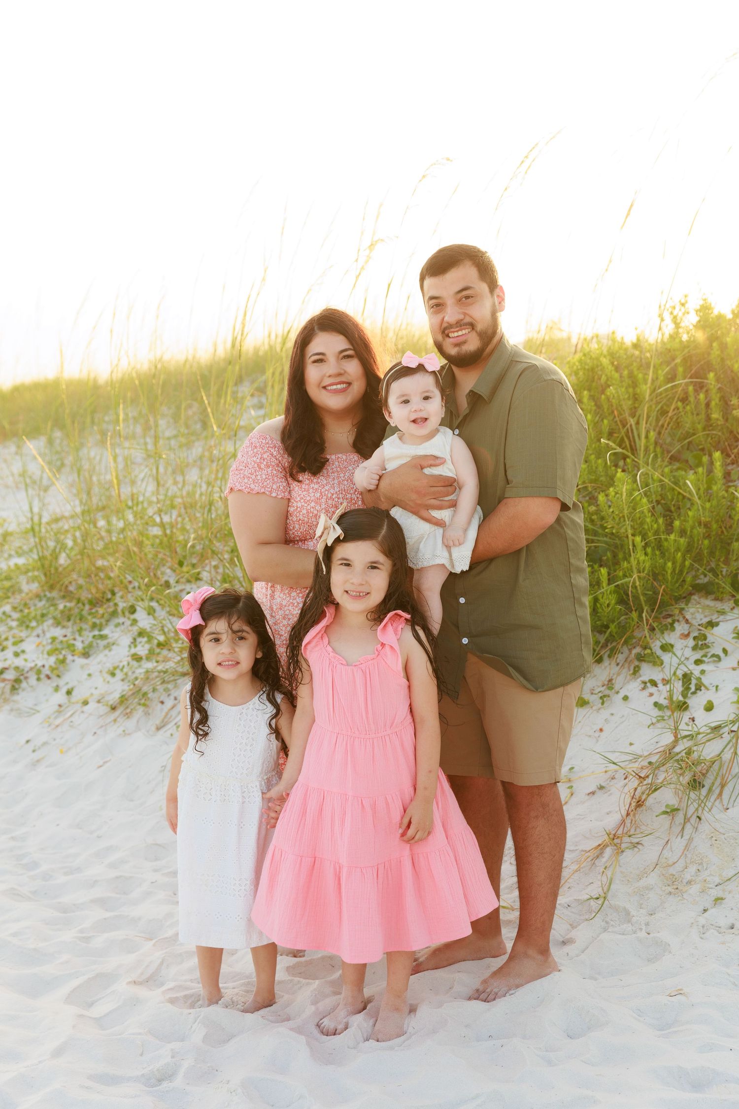 Family of five posing on sandy beach at sunset, parents with three young daughters in pink and white dresses near sea oats.
