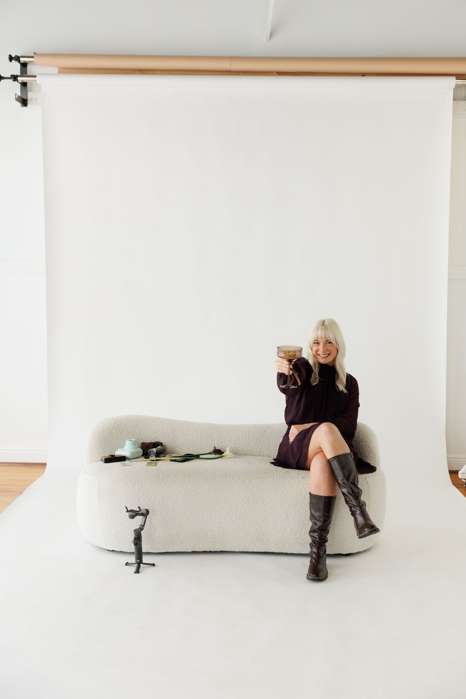 Person in black dress and boots lounging on white couch in photo studio setting with cocktail glass.