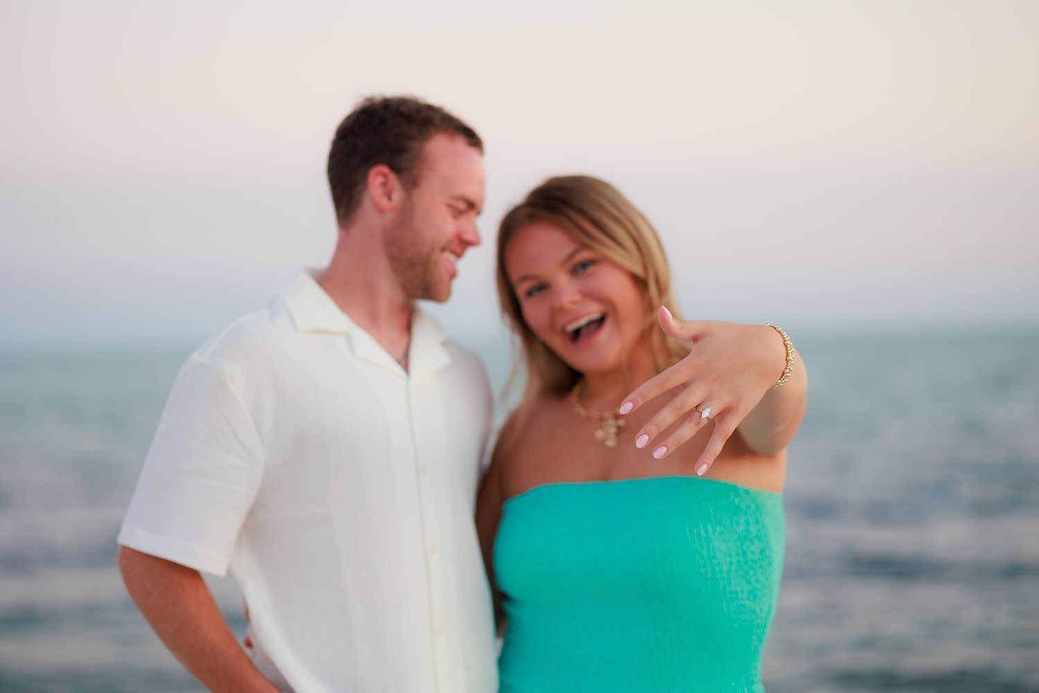 A happy couple in casual beachwear laughs together while embracing on a scenic oceanfront at sunset.