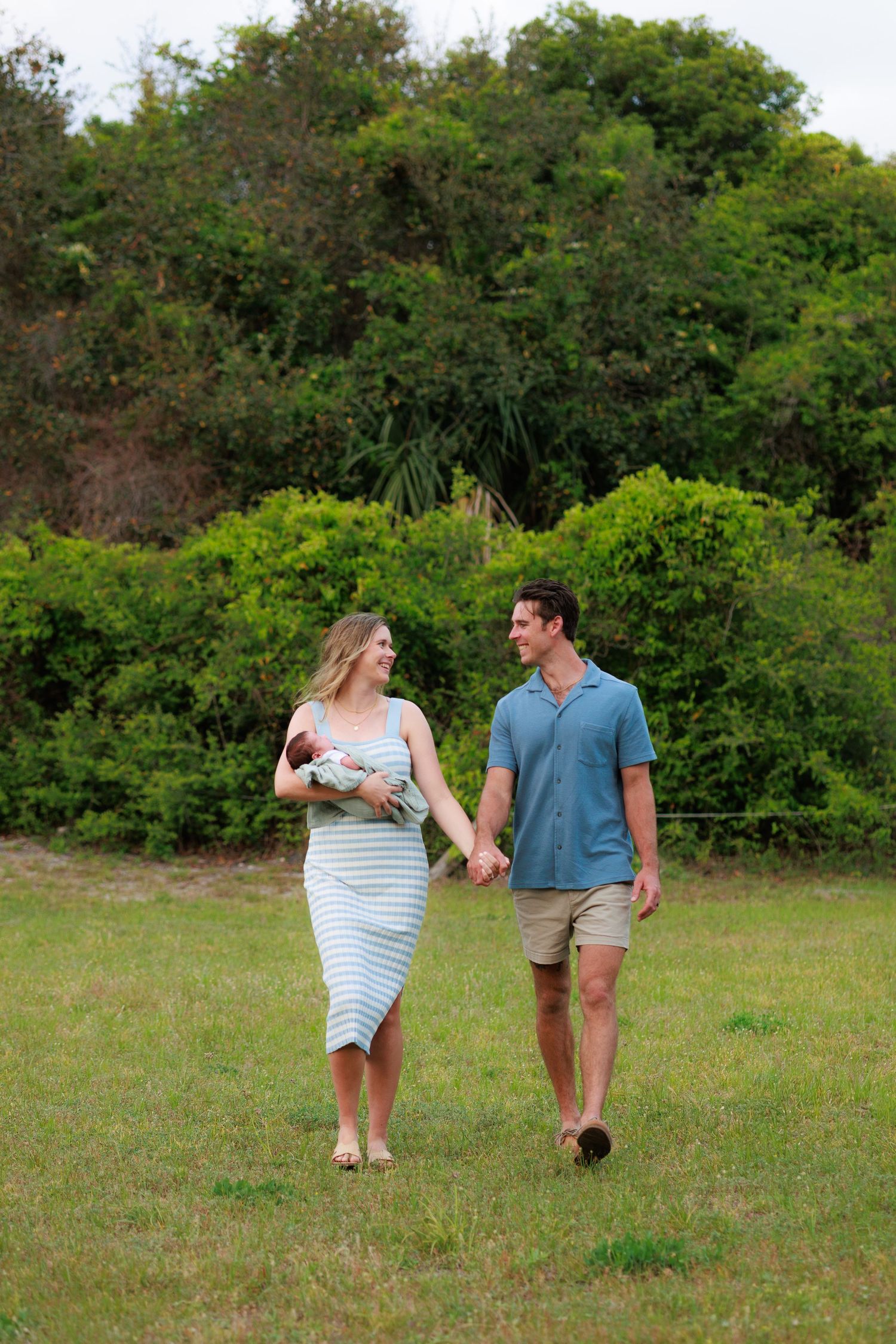 A couple walks together holding hands across a grassy field surrounded by lush green trees on a sunny day.