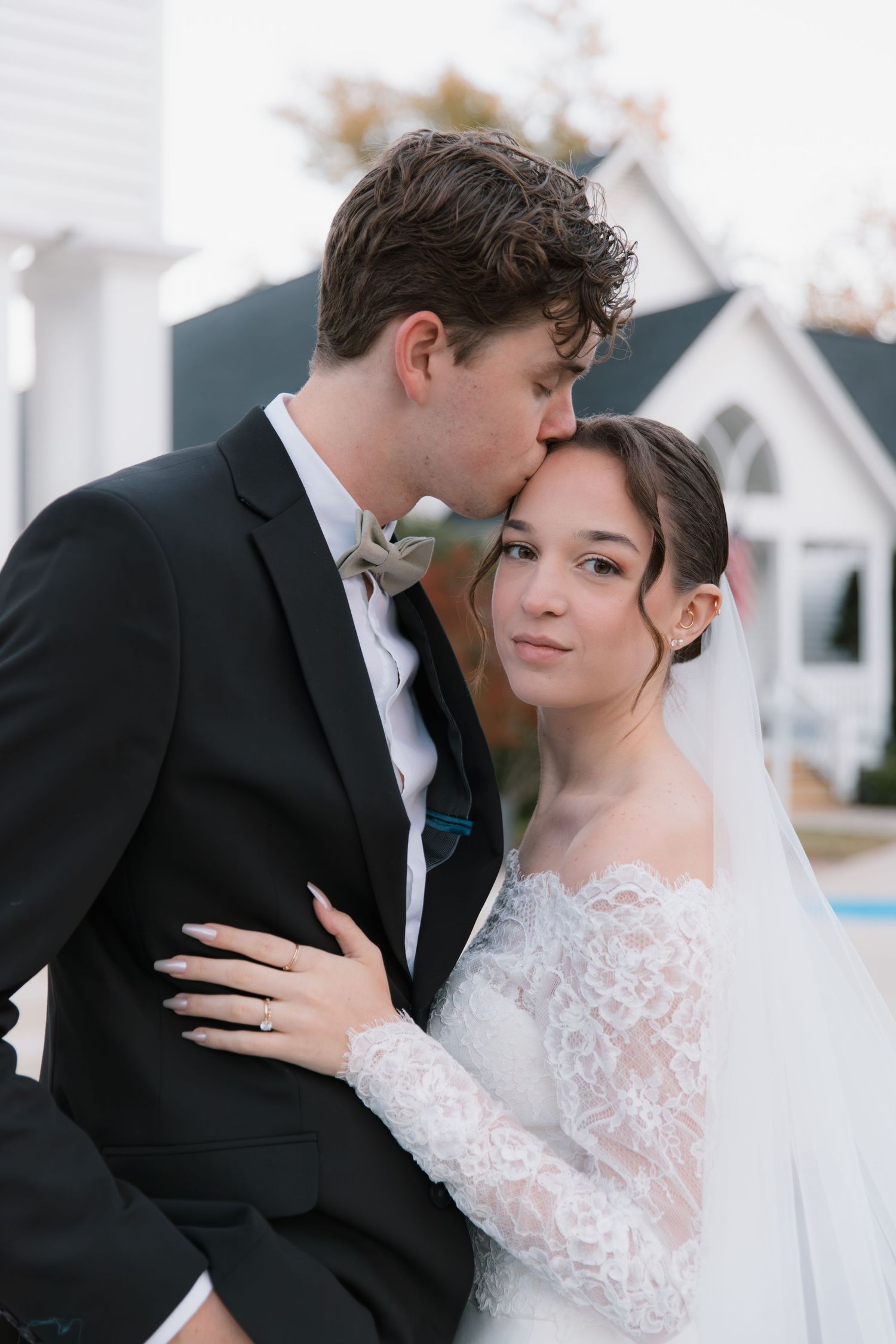 Romantic wedding couple embraces in front of white chapel with elegant lace dress and black suit details.