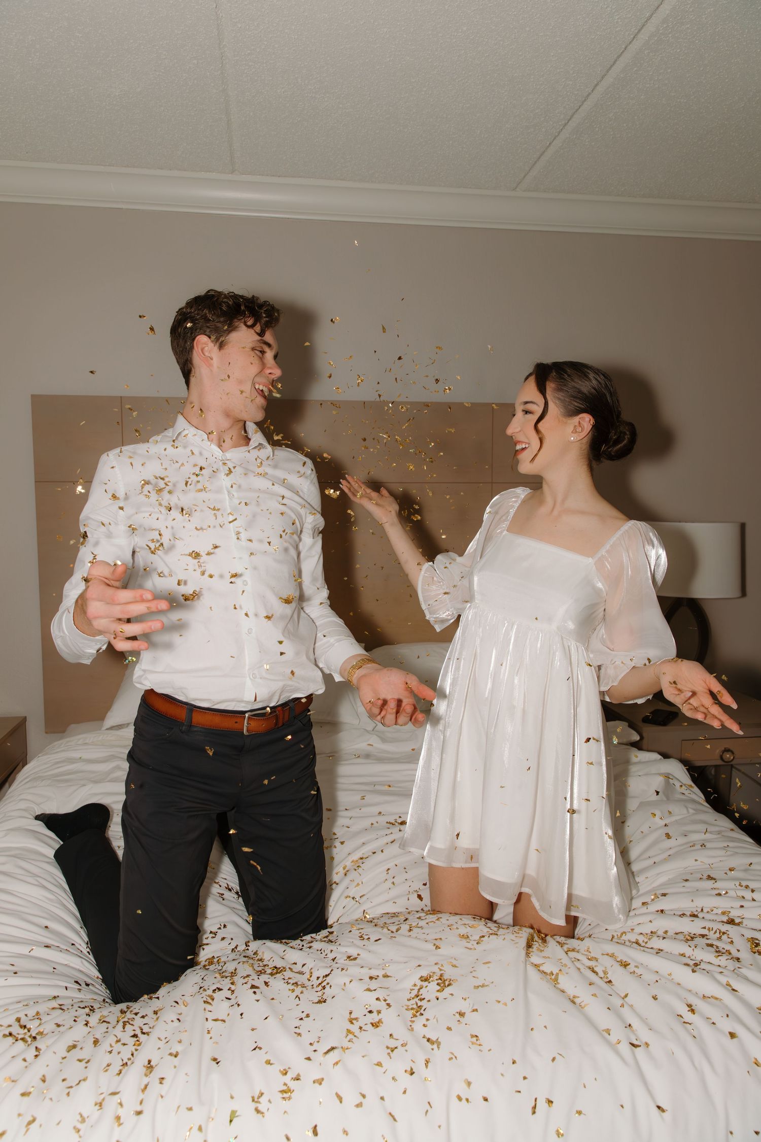 Playful couple having fun on a bed throwing confetti and laughing in wedding attire.