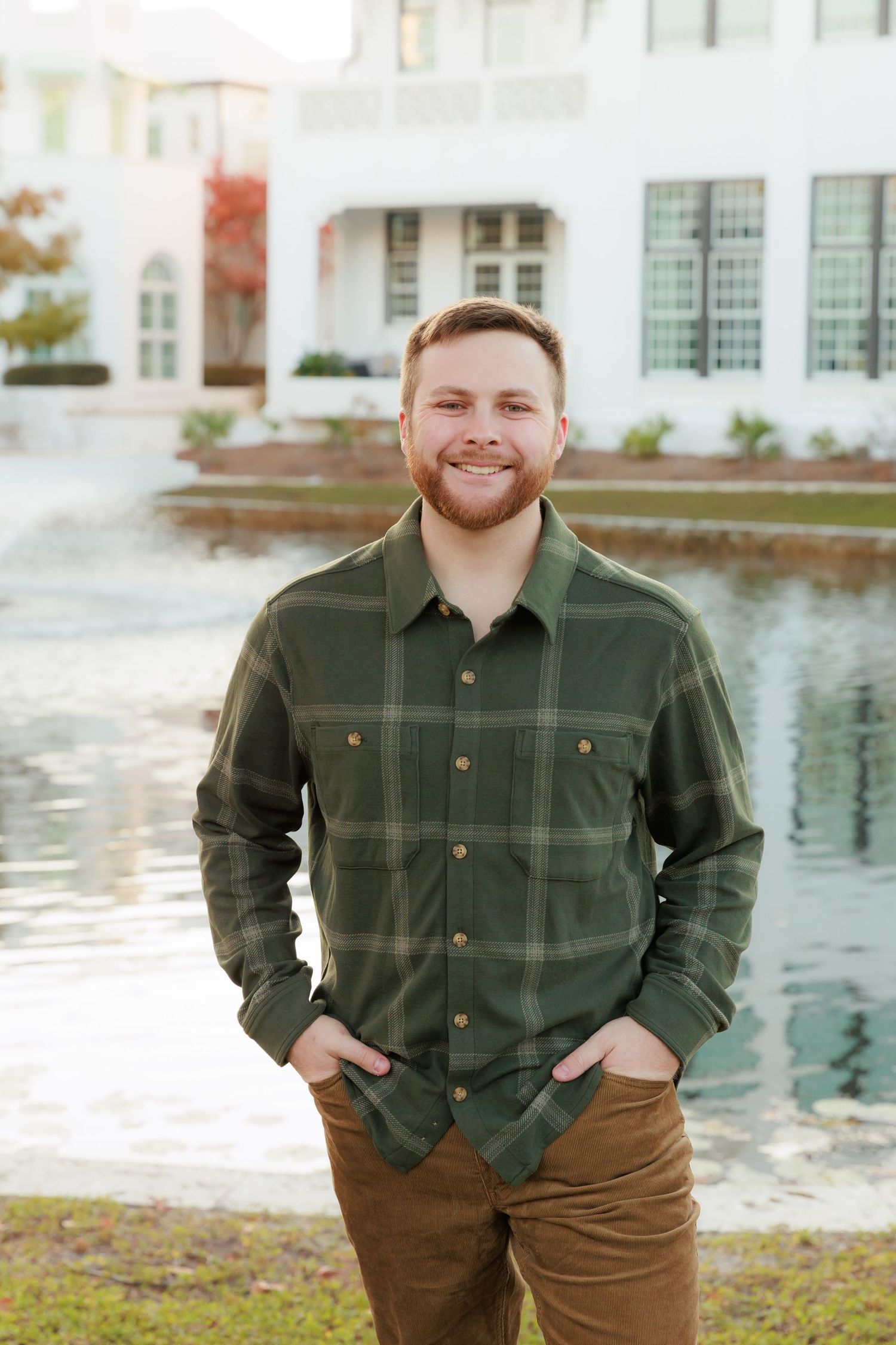 Person in green plaid shirt and brown pants stands smiling by pond with white building in background.