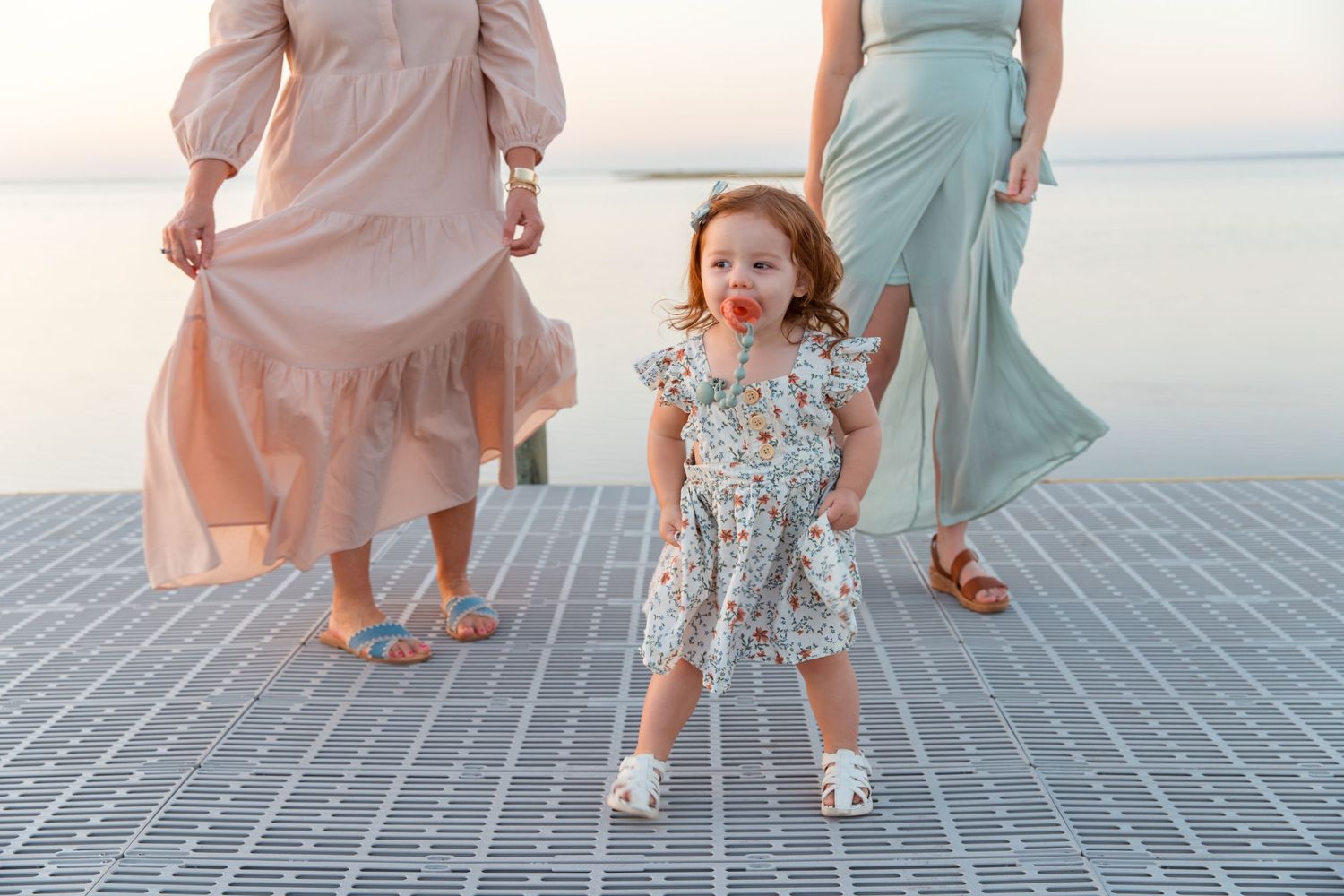 People in pastel dresses walking and playing on a wooden dock by the water at sunset.