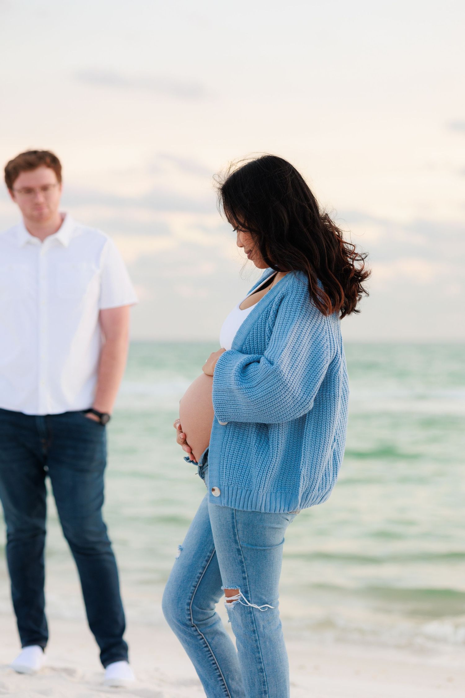 Expectant couple standing on beach at sunset with woman in blue sweater and ripped jeans.
