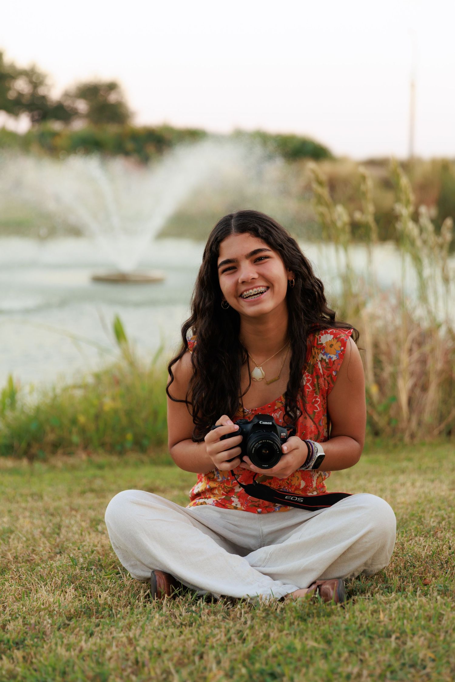 Photographer sits cross-legged on grass holding camera with fountain and lake in background.
