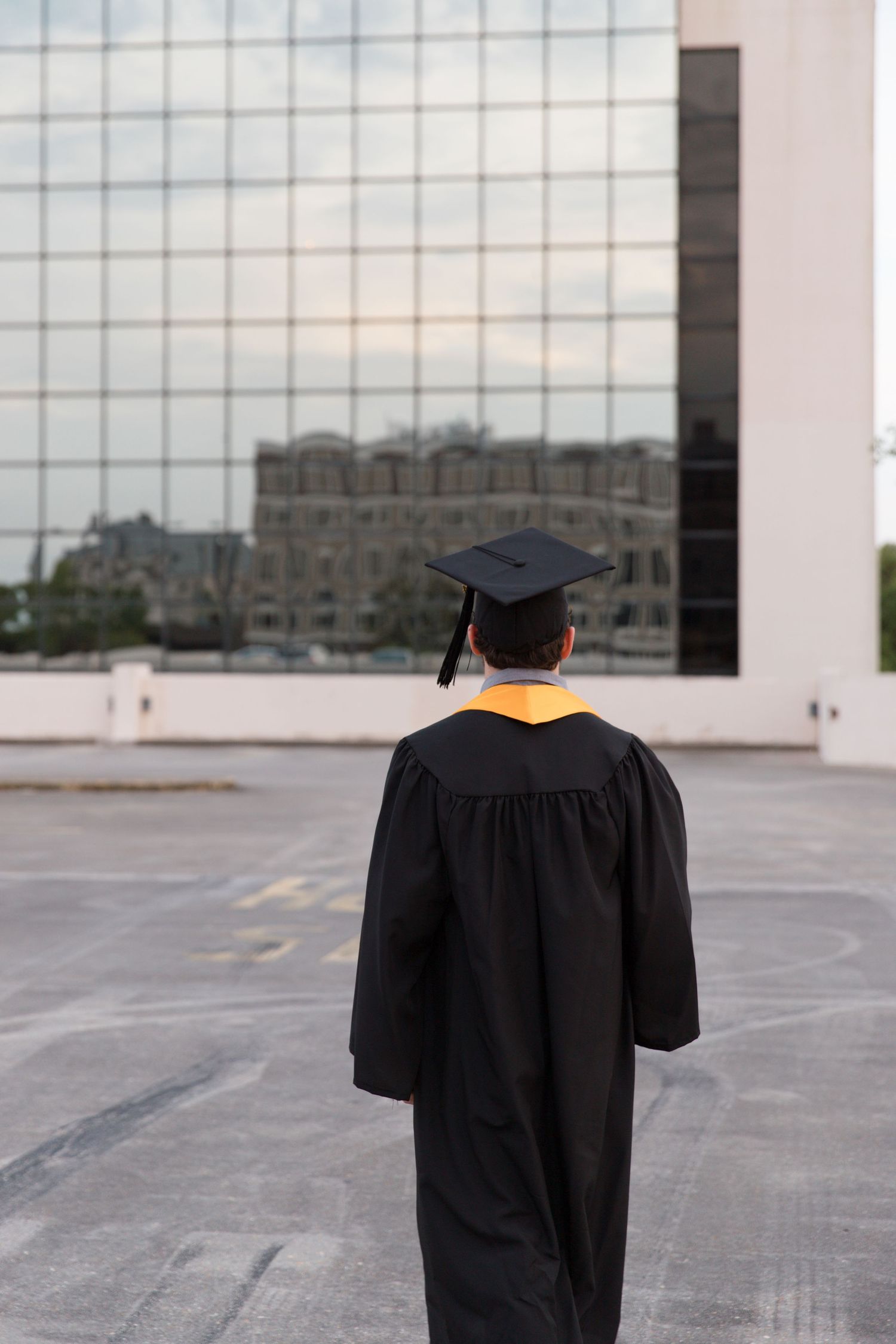 Graduate in black cap and gown standing on rooftop facing glass building.