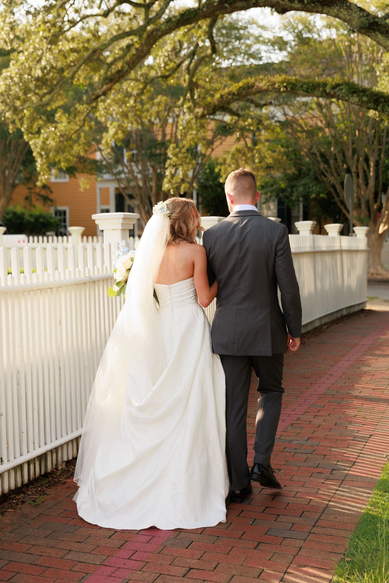 Newlyweds walk along brick path beside white picket fence under tree canopy.