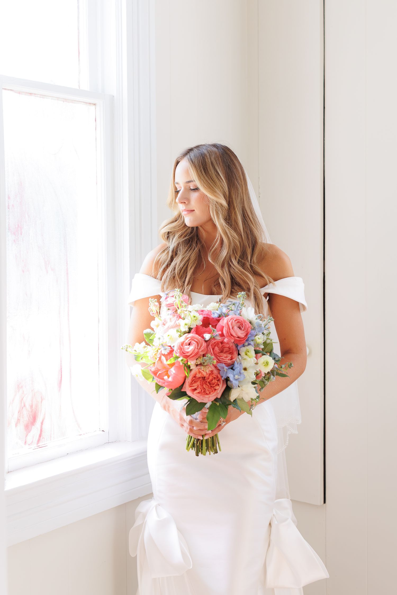 Bride in an off-shoulder white wedding gown holds a romantic bouquet of pink and white flowers by a bright window.