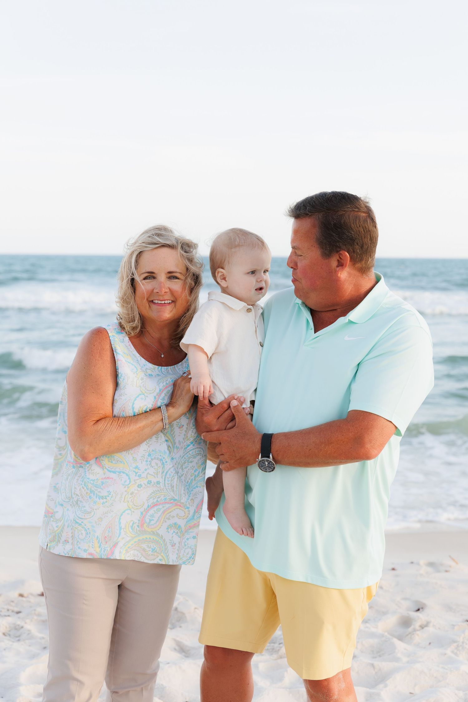 Grandparents smiling and holding baby on sunny beach with ocean waves in background.