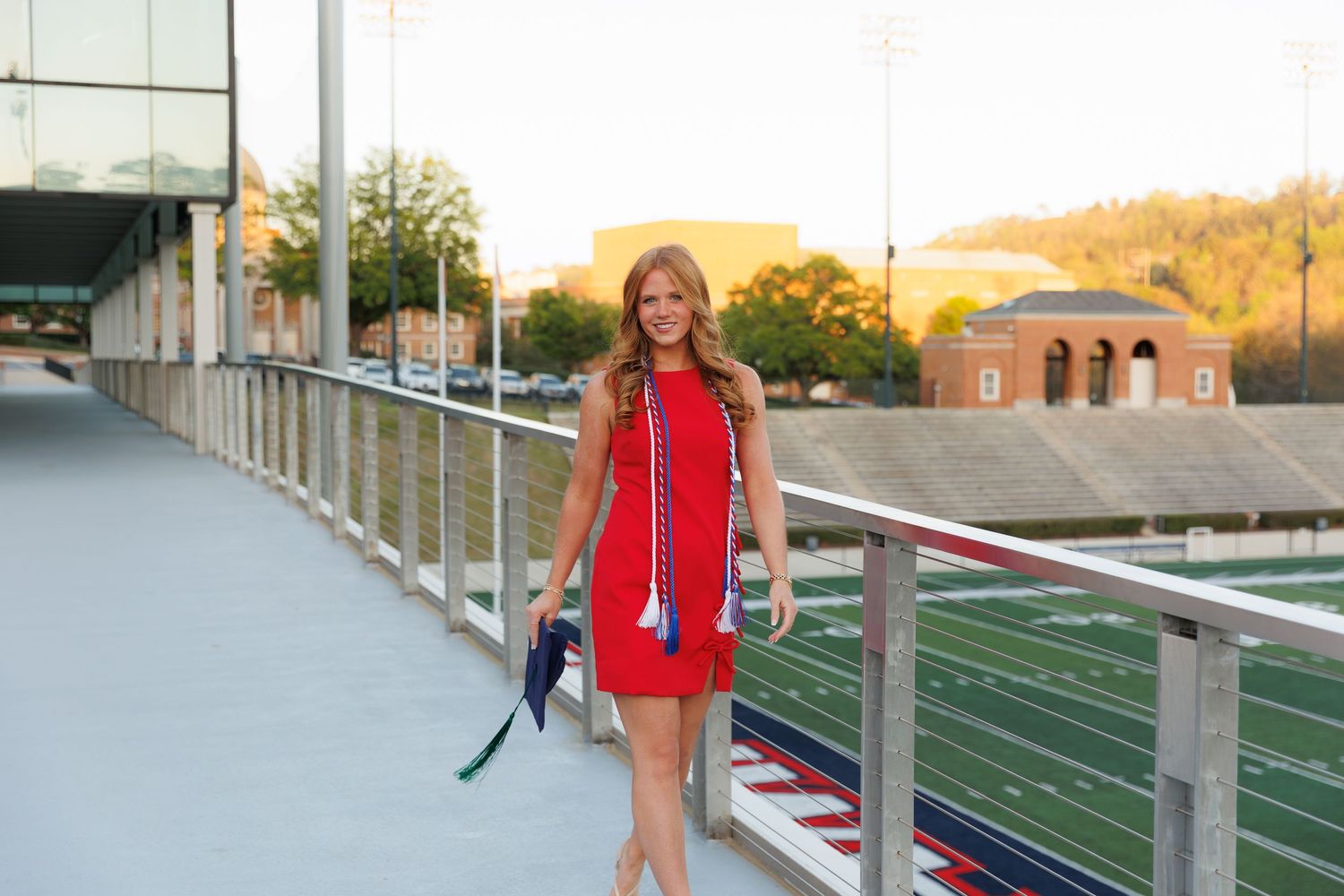 Young woman in red dress and graduation tassel walks on stadium bridge at sunset.