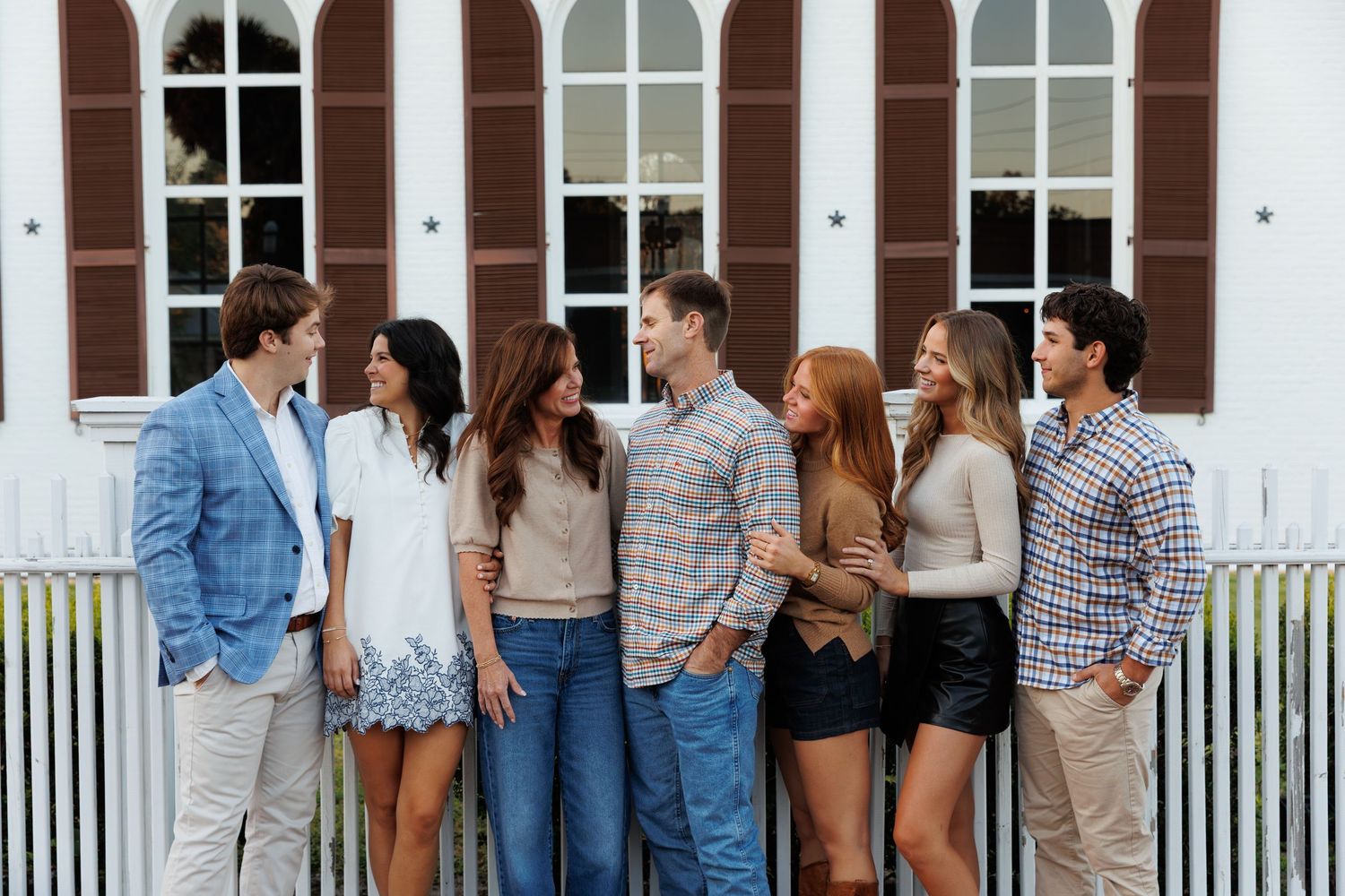Group of six young adults posing together outdoors near a white fence and building with shuttered windows.