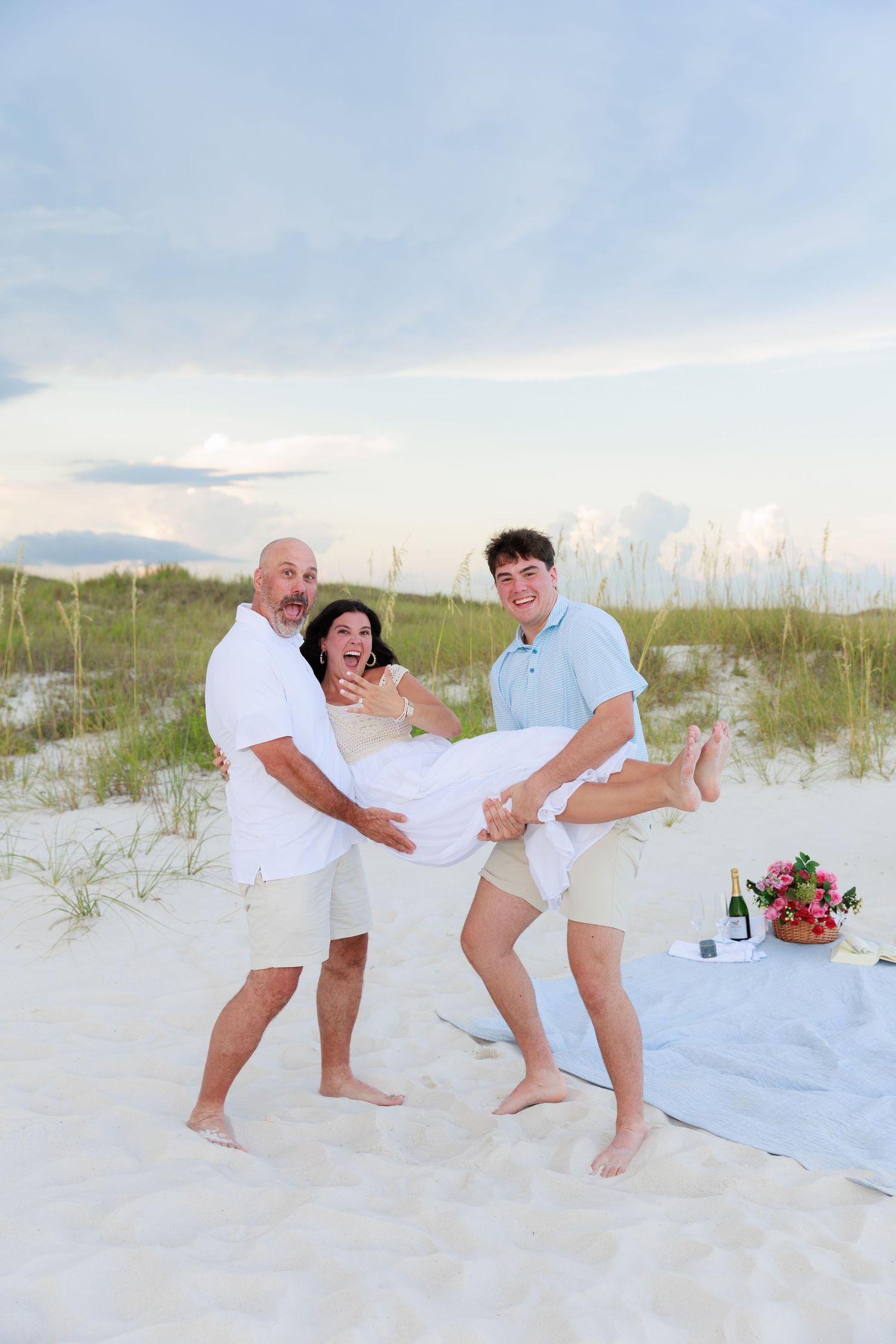 A joyful group in white outfits poses playfully on a sandy beach with sea grass and cloudy skies in the background.