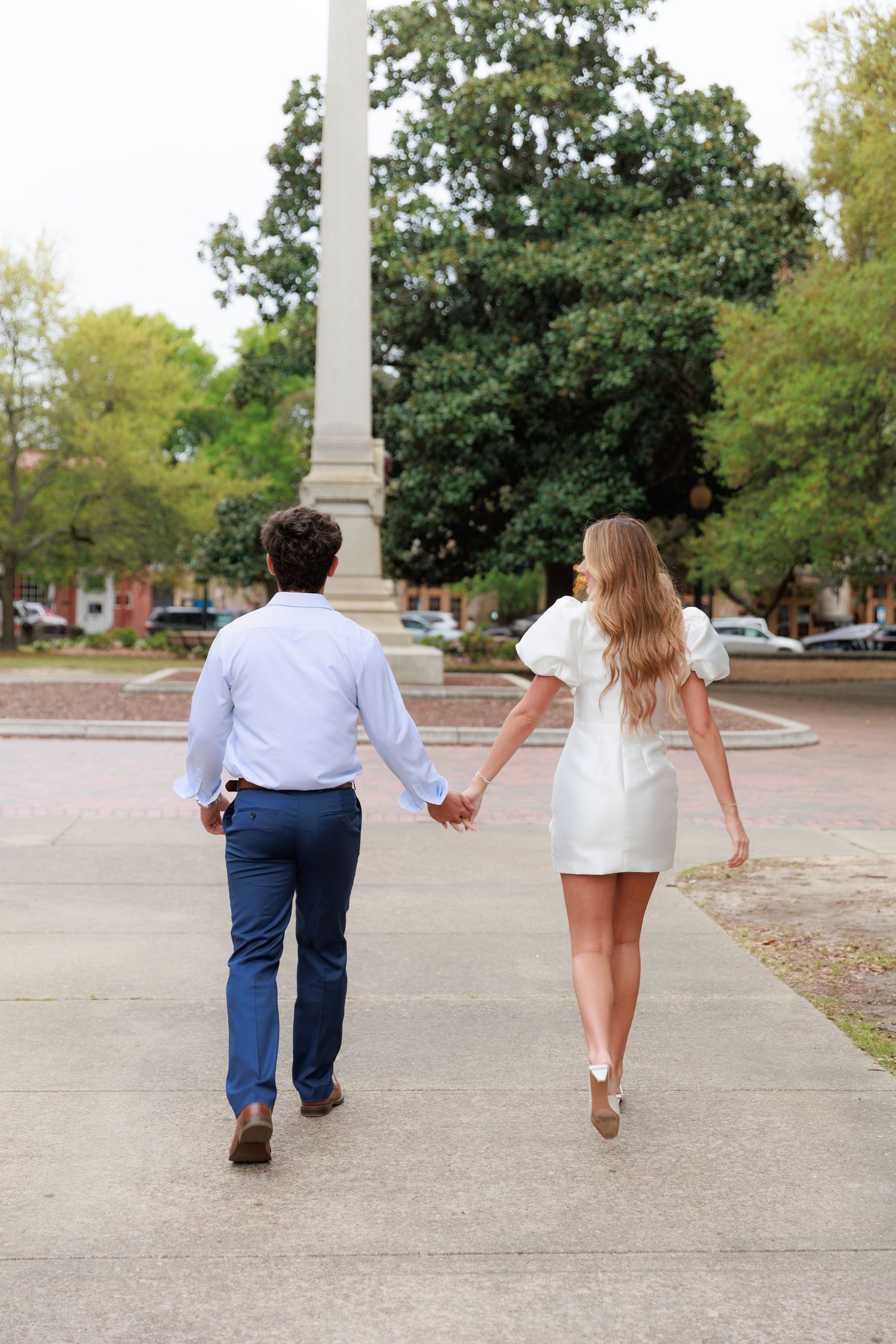 A couple holding hands while walking away from the camera on a paved path surrounded by green trees.
