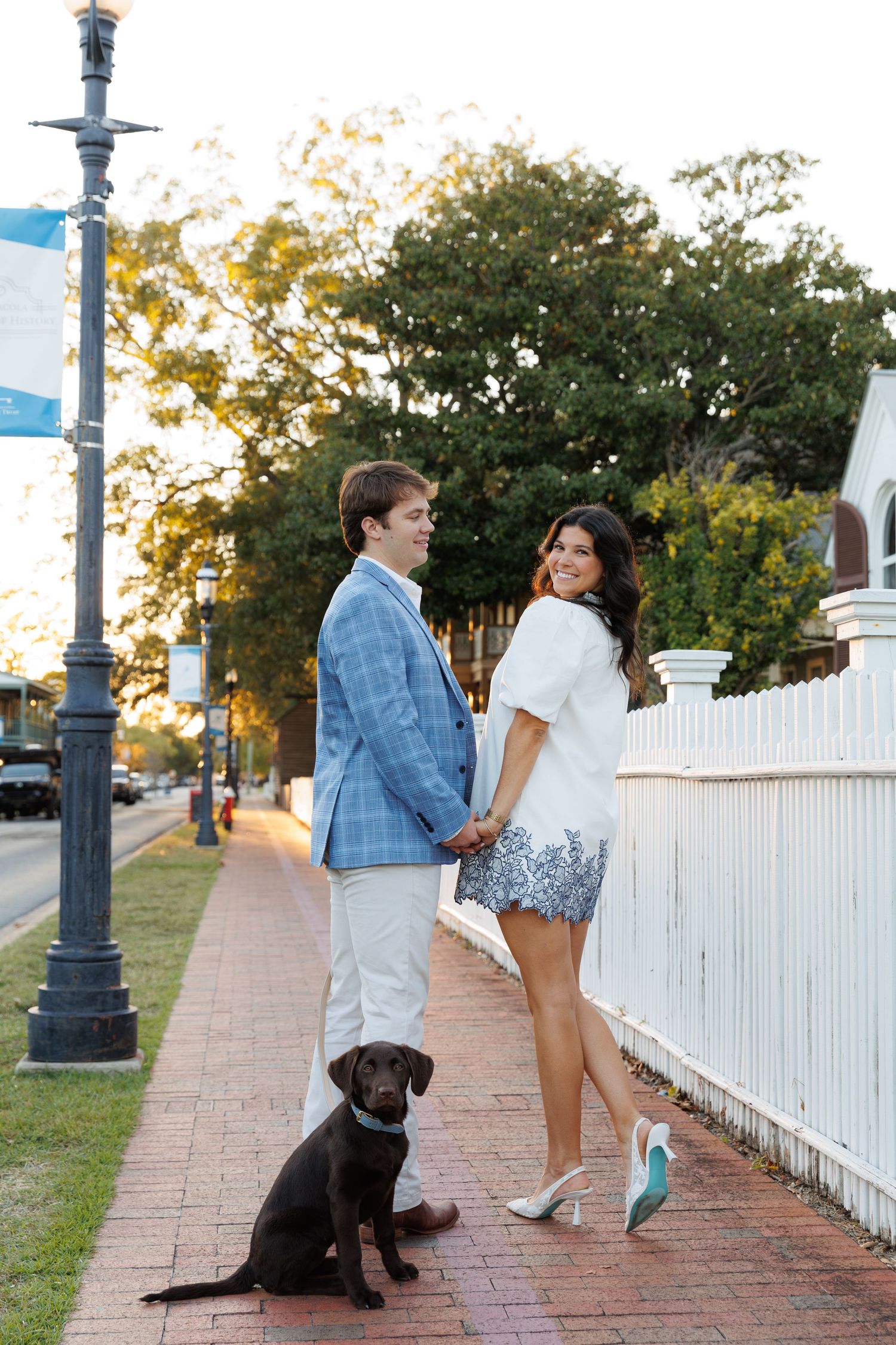 A couple and their black dog stand along a white picket fence on a brick sidewalk at sunset.