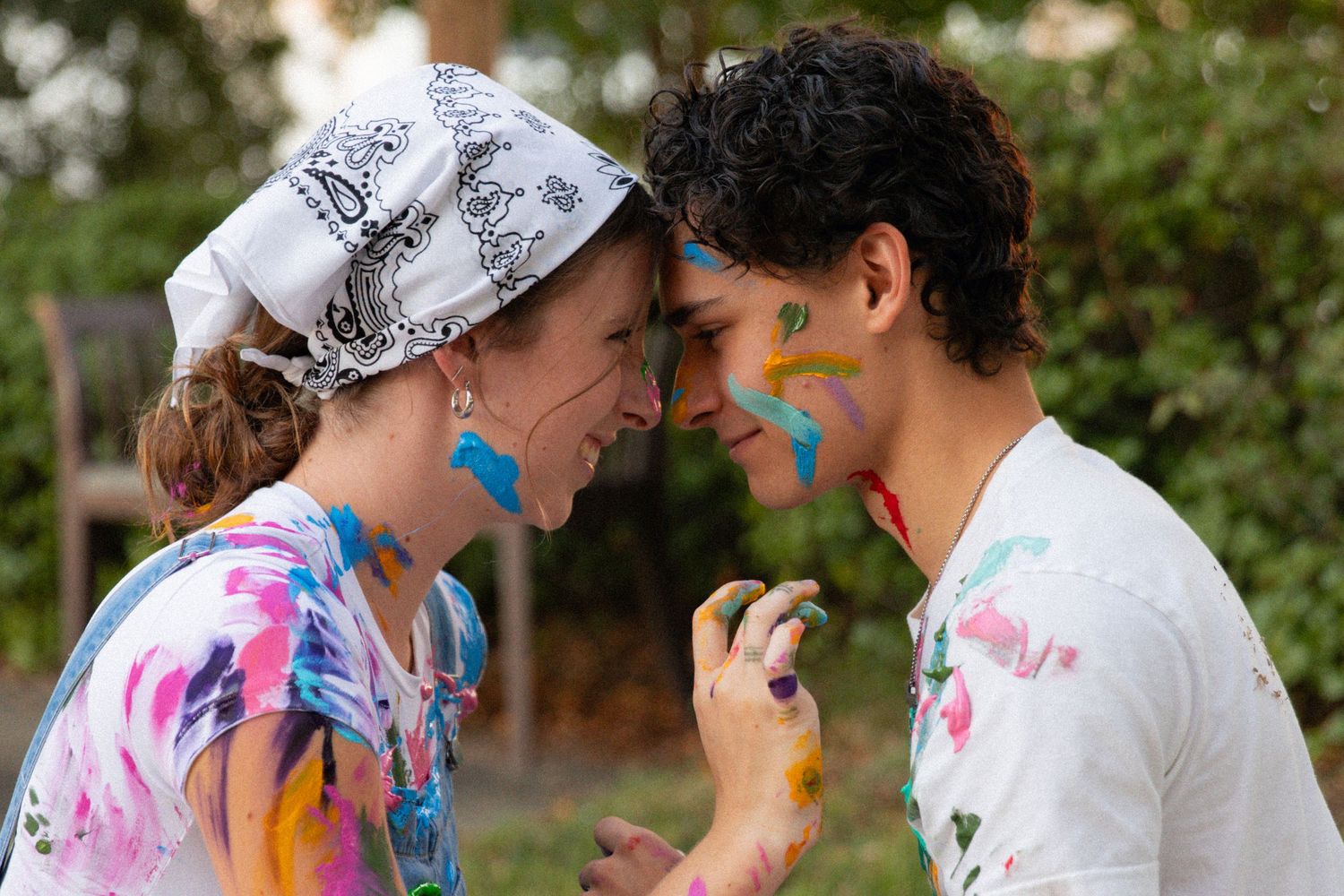Two people with colorful face paint wearing tie-dye and white shirts lean in close while smiling outdoors.