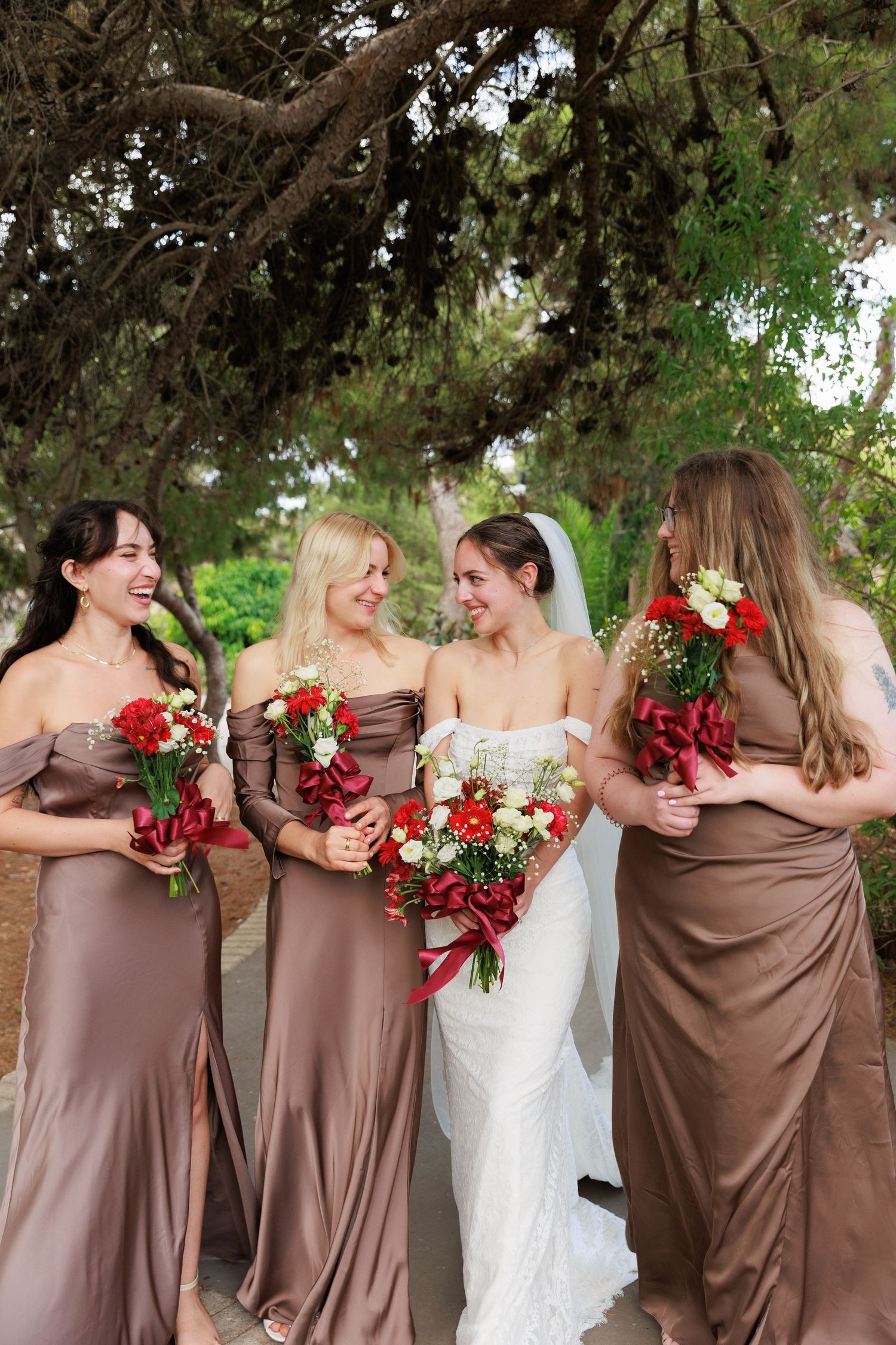 Bridesmaids in chocolate brown dresses pose together with red bouquets under a tree at an outdoor wedding.