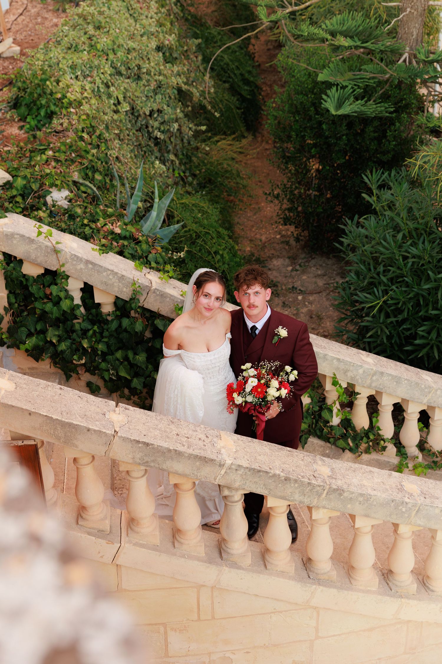 A couple stands along a stone balustrade in a garden setting with the bride in white and groom in burgundy.