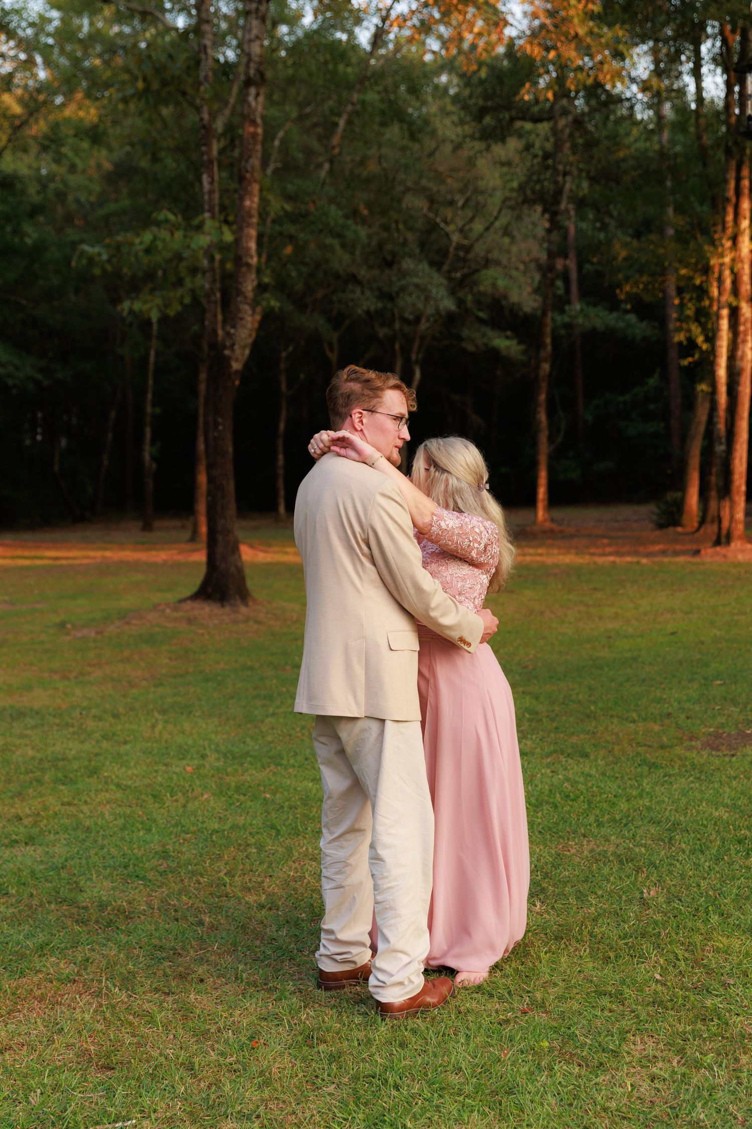 A couple shares a romantic moment at sunset in a grassy park with tall trees in the background.